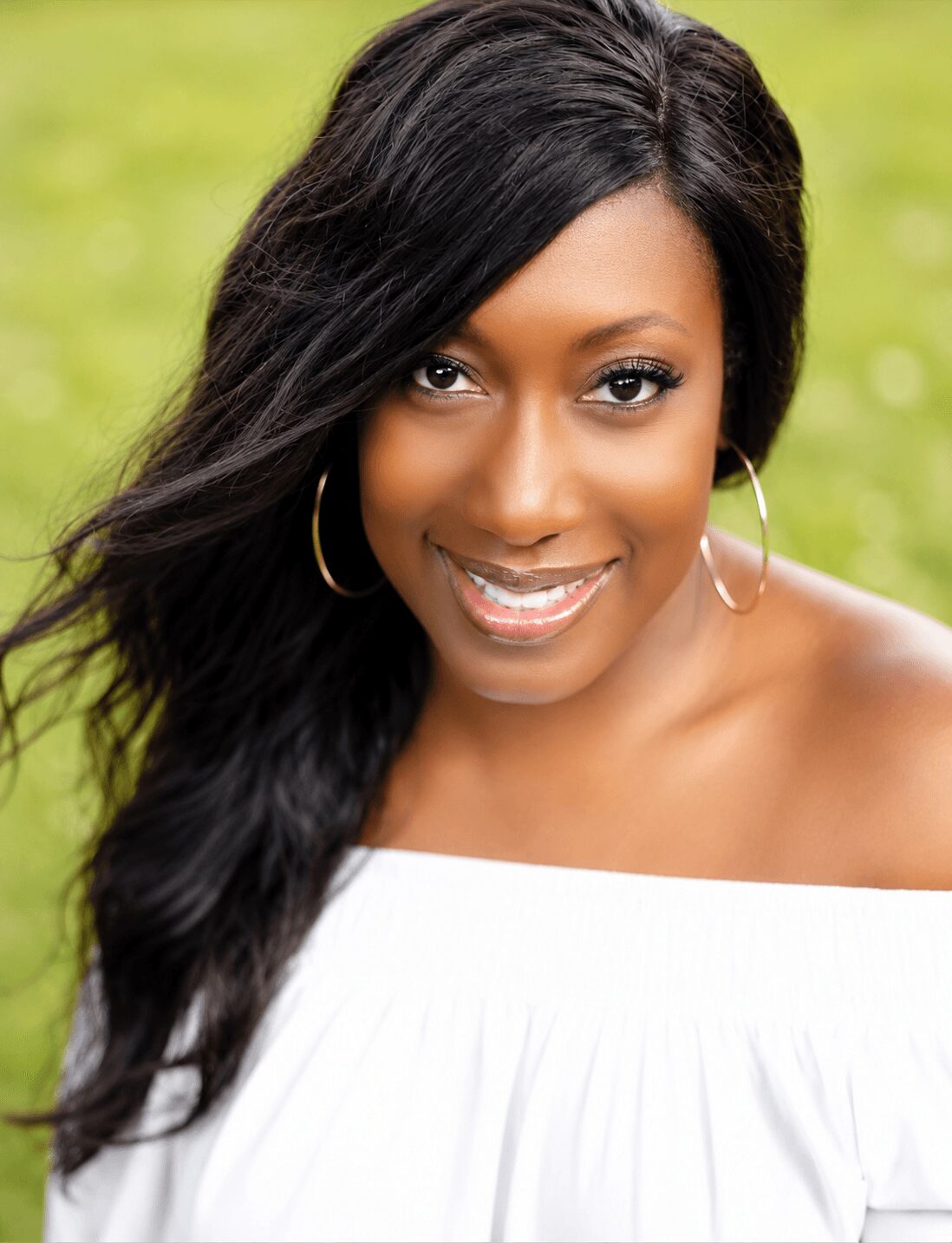 Woman with long black hair, smiling, wearing hoop earrings and a white off-shoulder top outdoors.