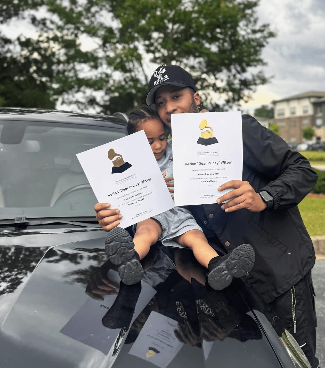 Person holding a certificate and a child sitting on a car hood outdoors with trees and buildings in background.