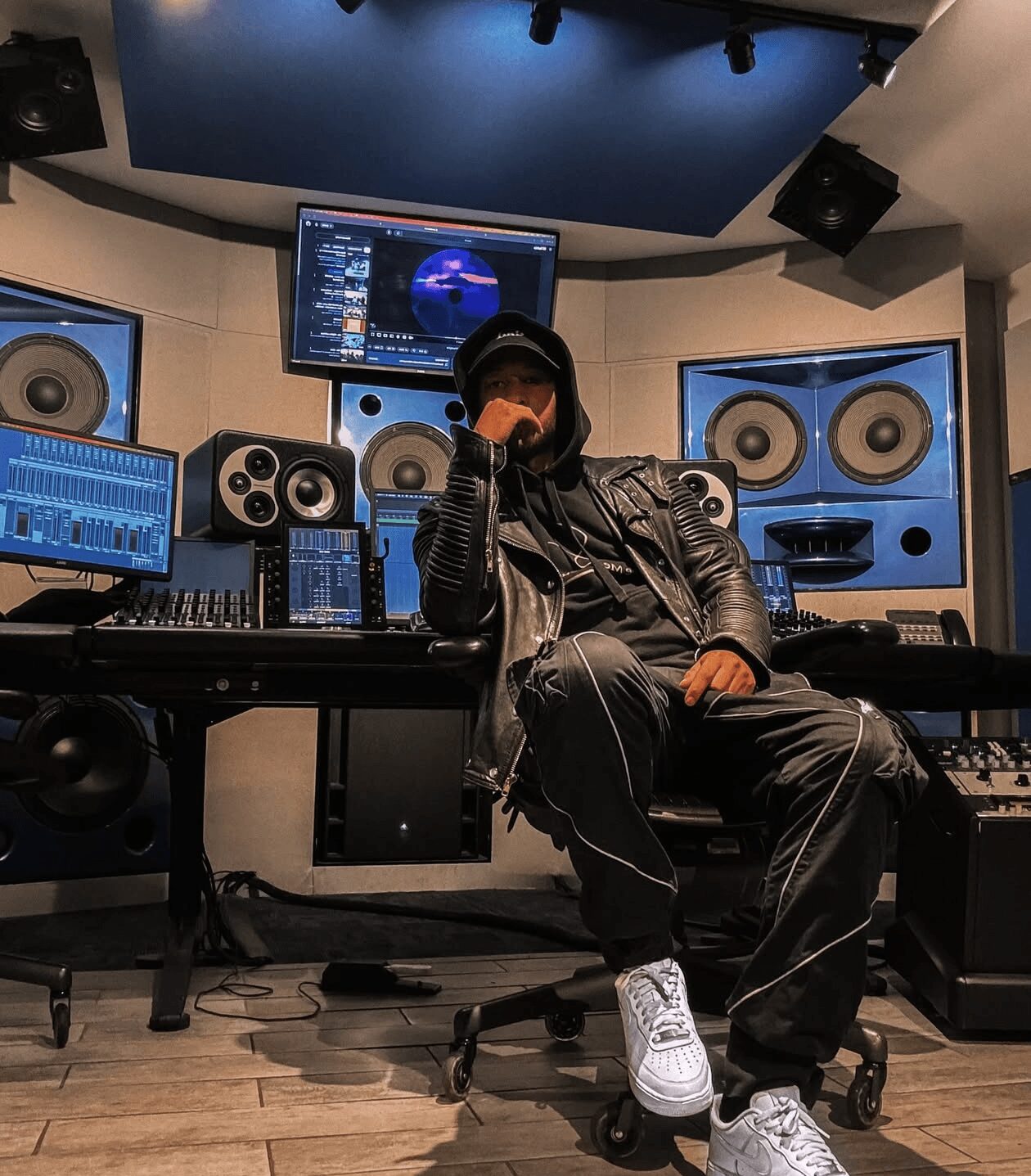 Person sitting in a music studio with speakers, mixing console, and computer screens, wearing a leather jacket and white sneakers.