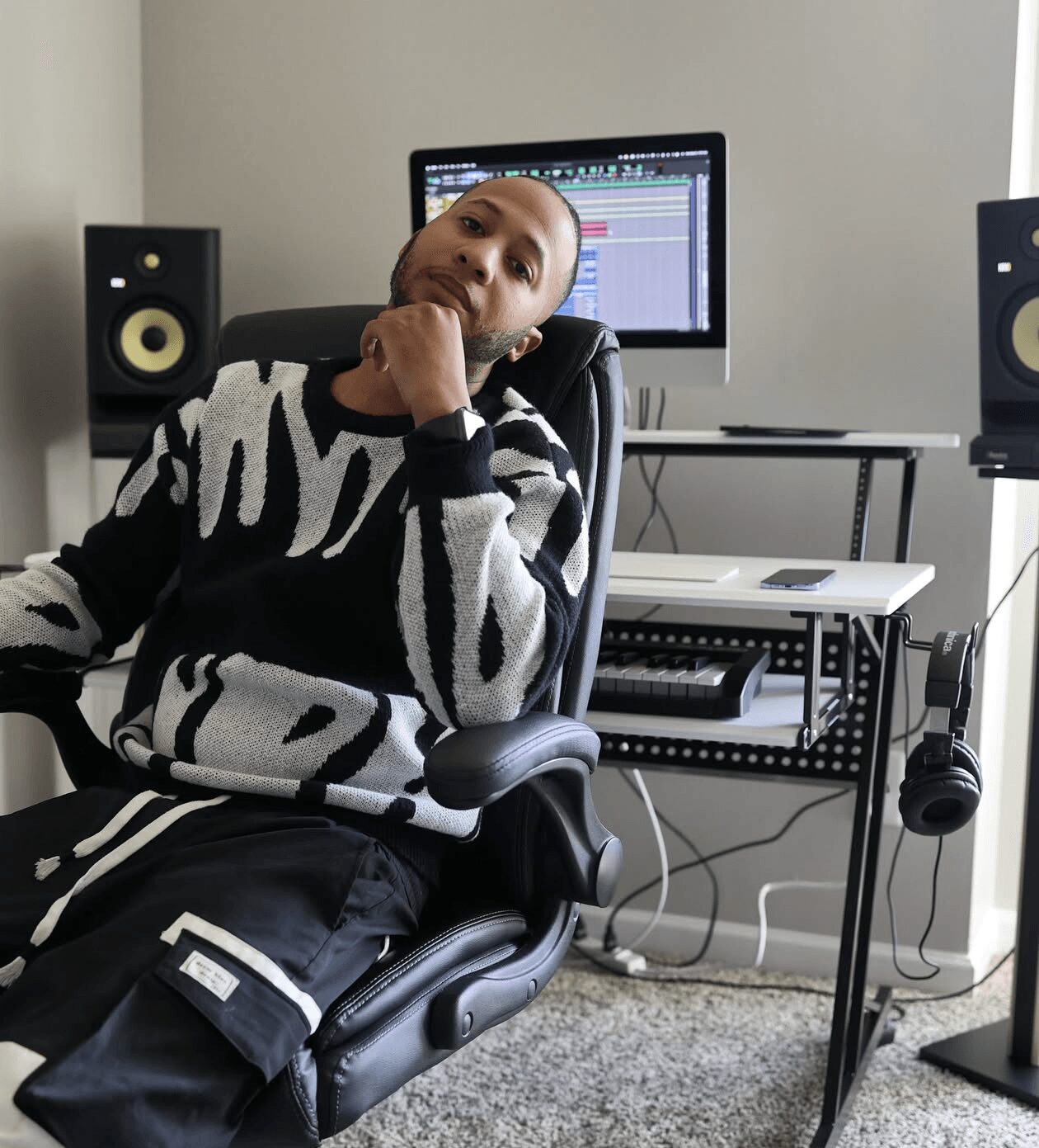 Person sitting in a chair in a music studio with computer and speakers, resting chin on hand, looking at camera.