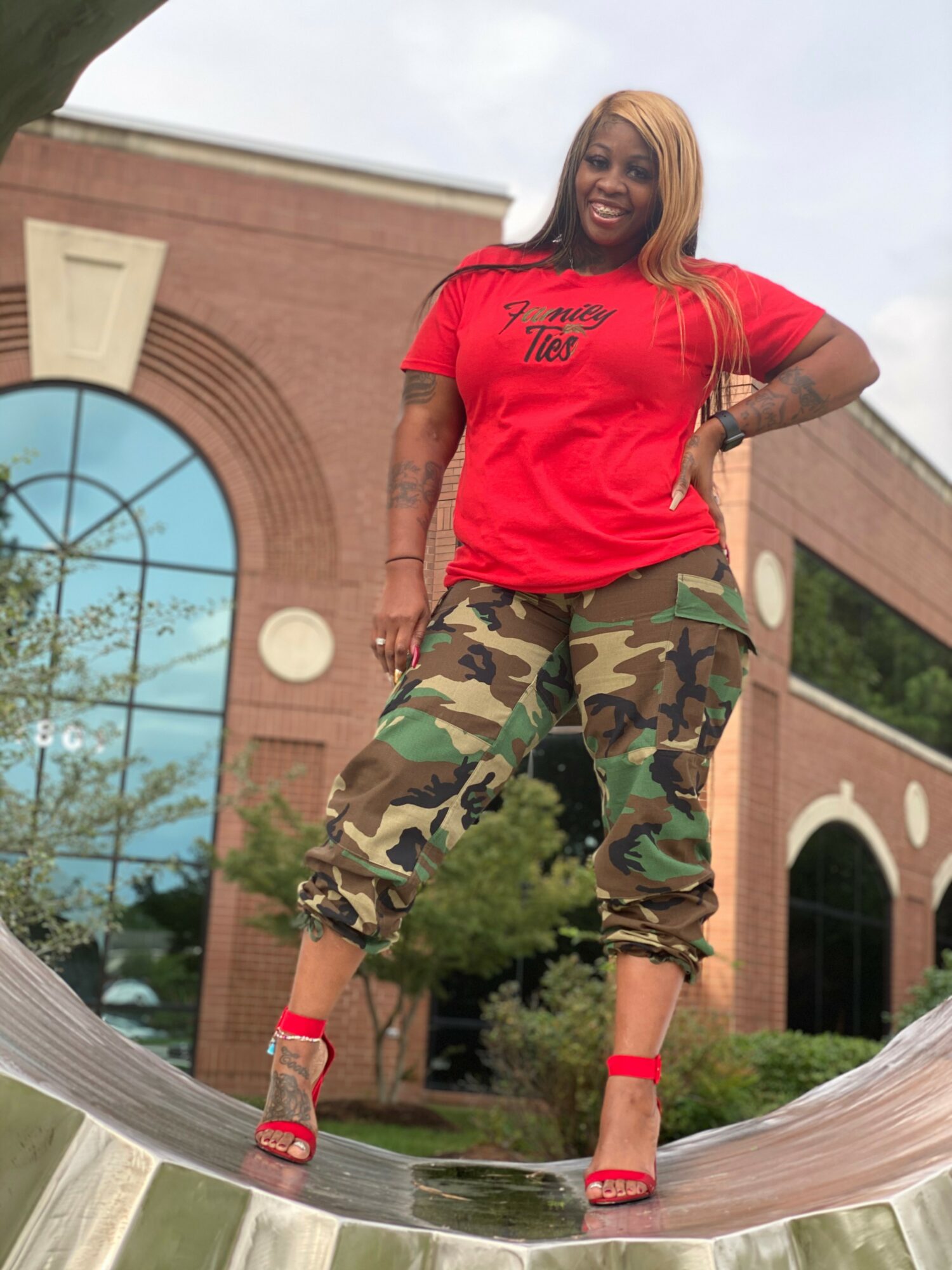 Woman with long hair in red t-shirt and camouflage pants standing on a curved metallic surface outdoors, smiling.