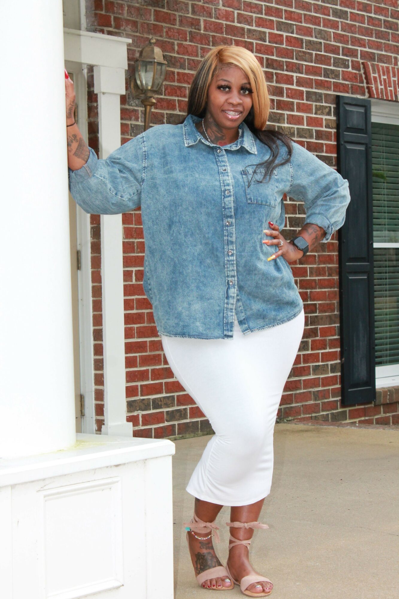 Woman standing in front of a brick wall, wearing a white dress, denim jacket, and high heels, smiling.