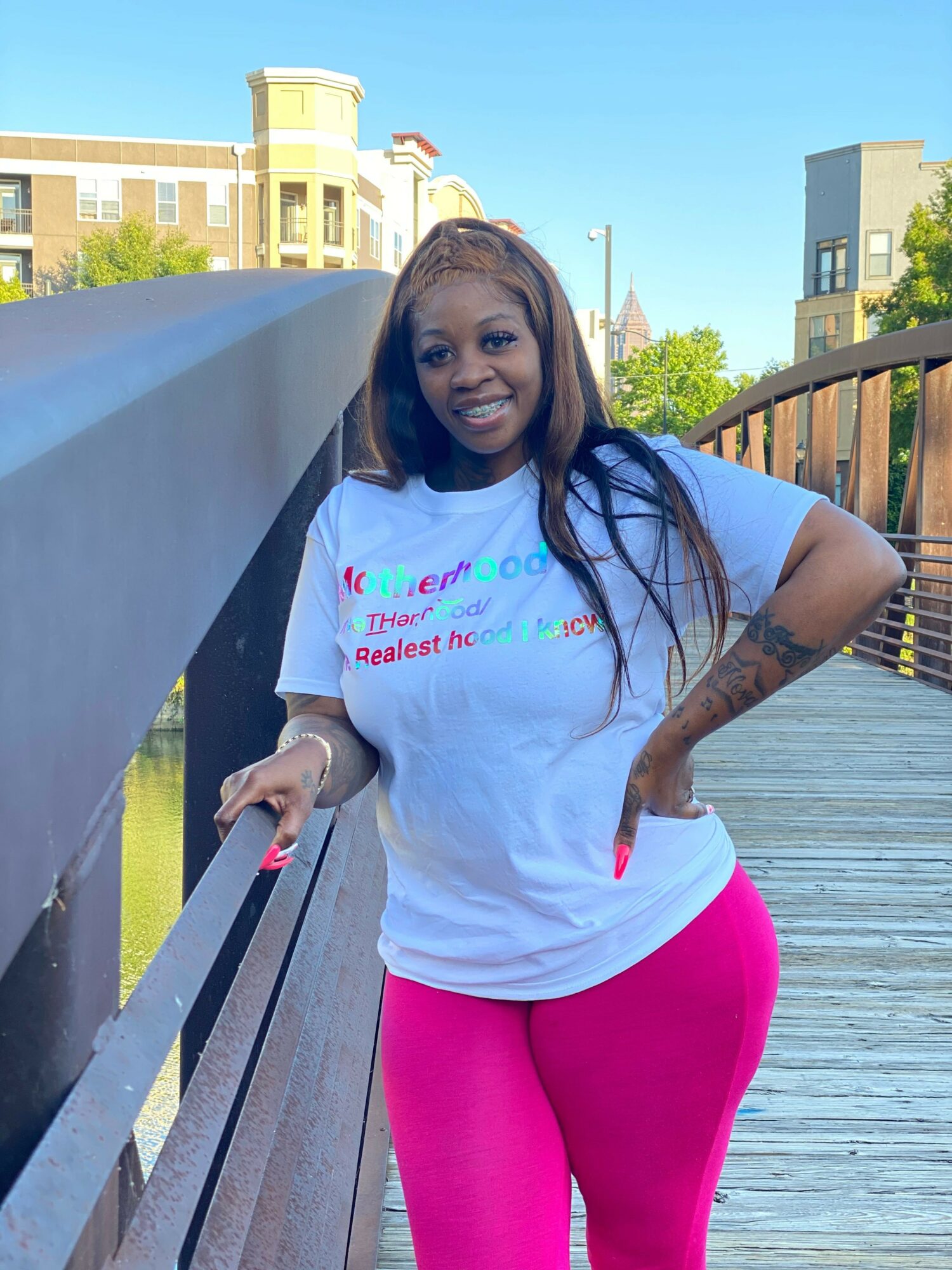 Woman with long dark hair, tattoos, wearing a white T-shirt and pink pants, standing on a wooden deck outdoors.