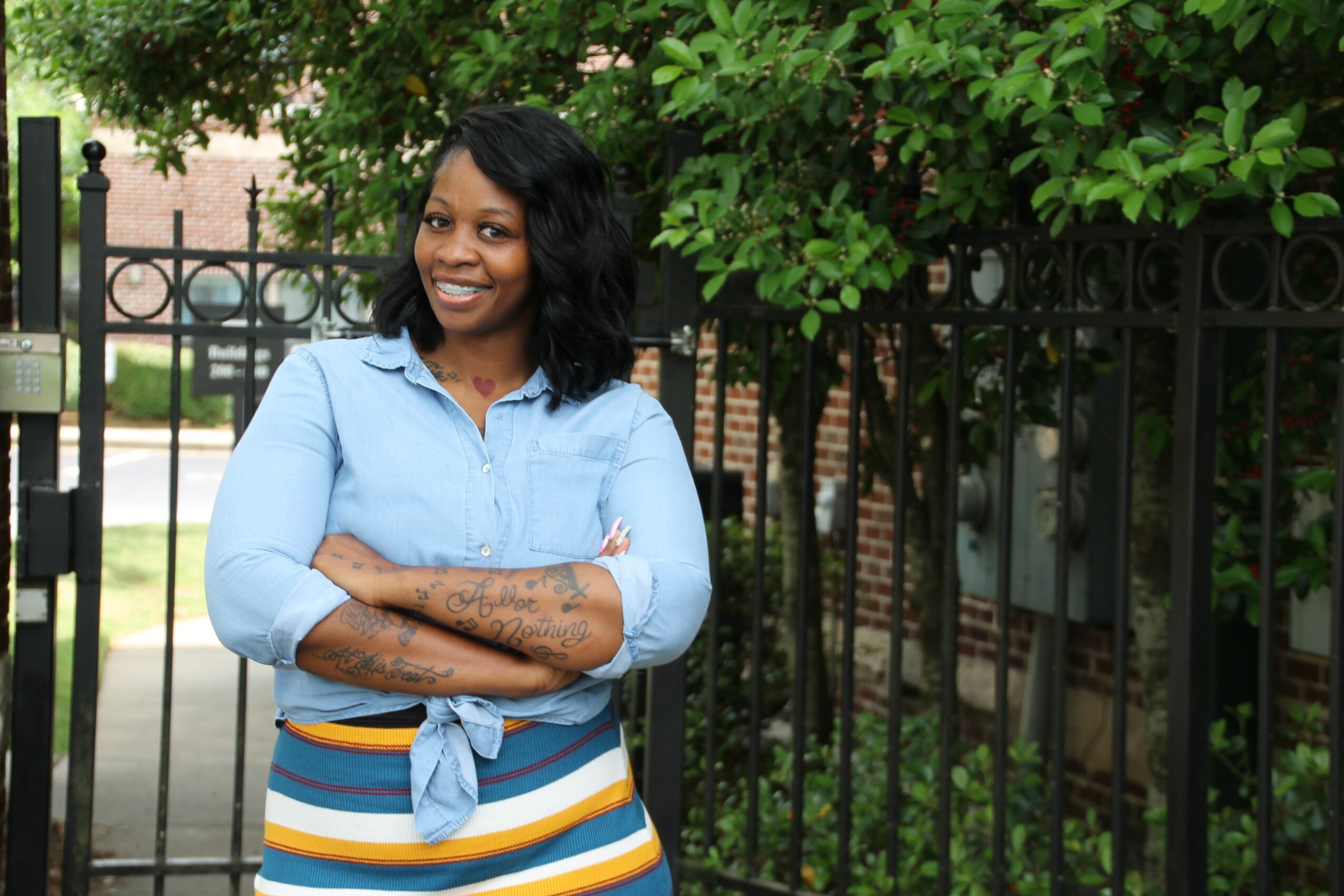 Woman with black hair smiling, wearing a light blue shirt and striped skirt, standing outdoors near a fence and trees.