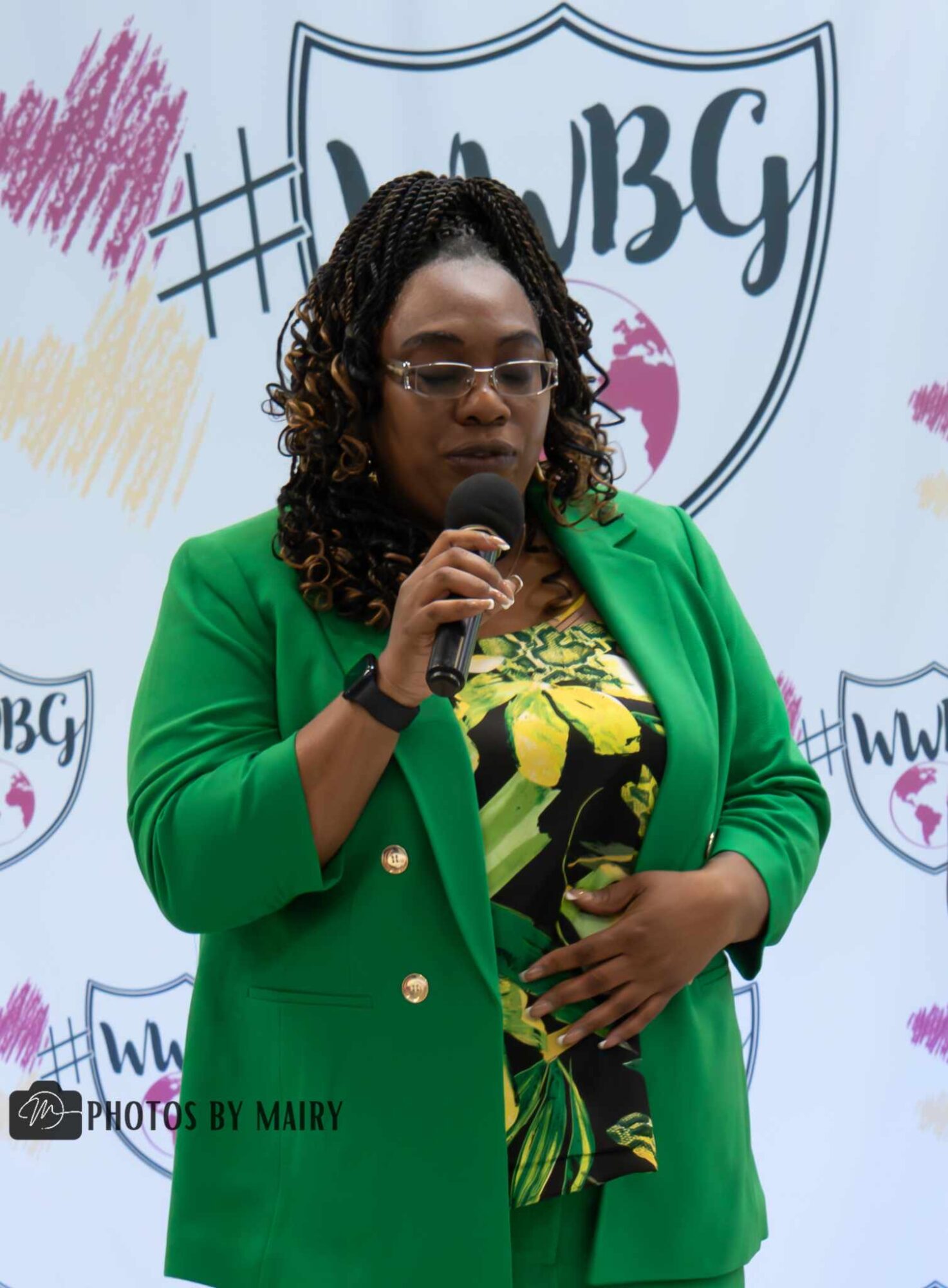 Woman in green blazer holding microphone, standing in front of a backdrop with logos and colorful designs.