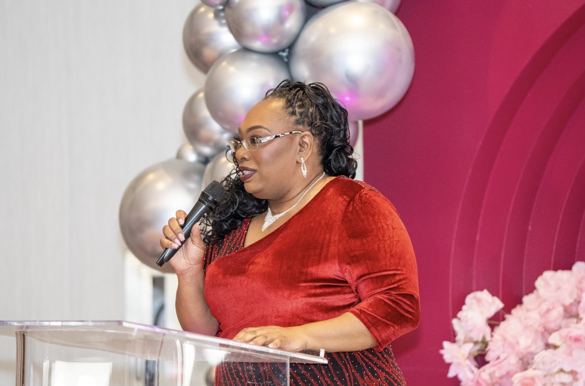 Woman in red dress speaking into a microphone at a decorated event with pink and silver balloons.