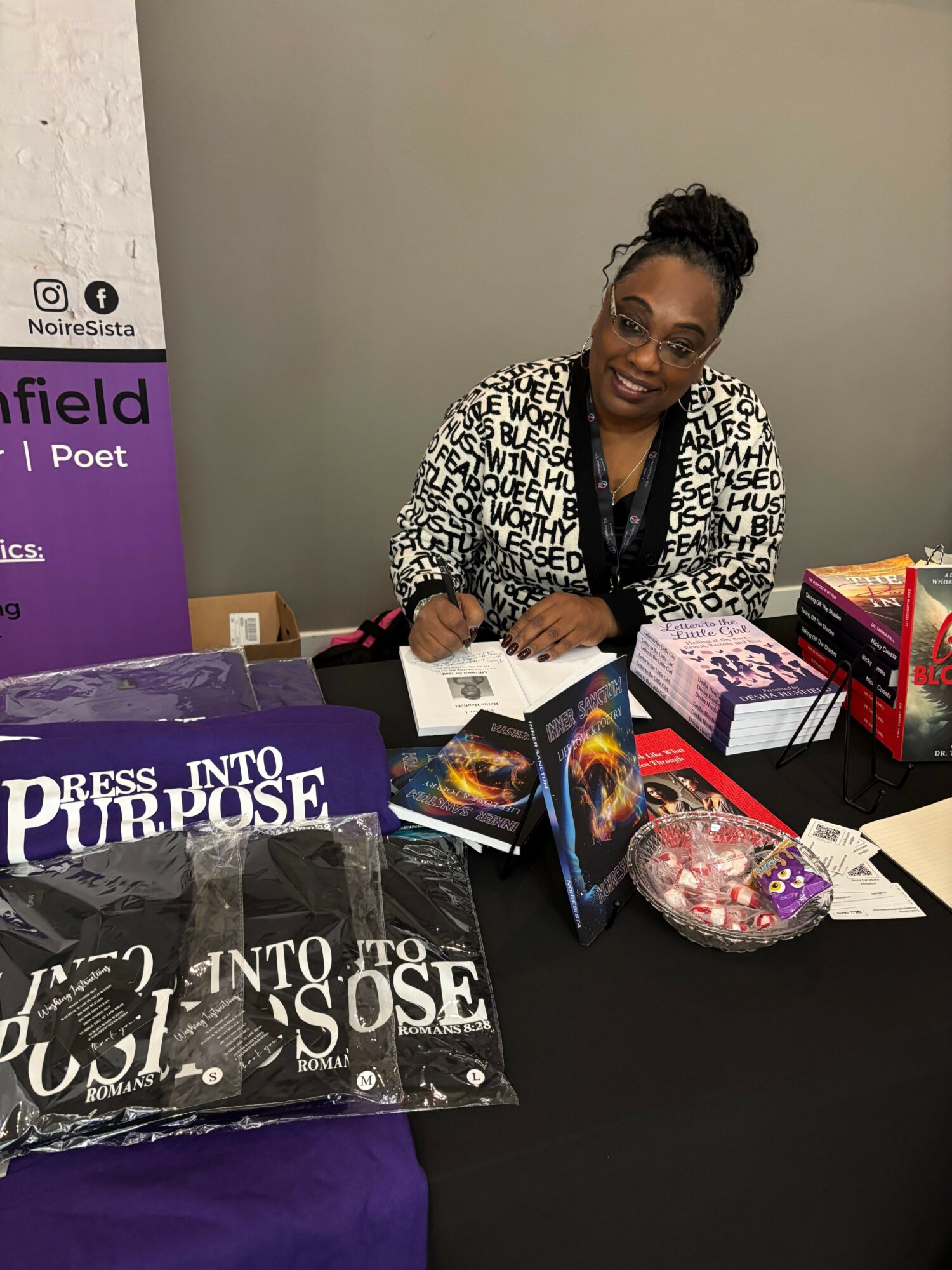 Woman with glasses and curly hair at a table with books, a purple banner, and a bowl of candies.