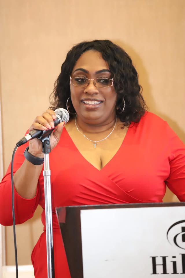 Woman with curly hair and glasses speaking into a microphone, wearing a red top, standing behind a podium.