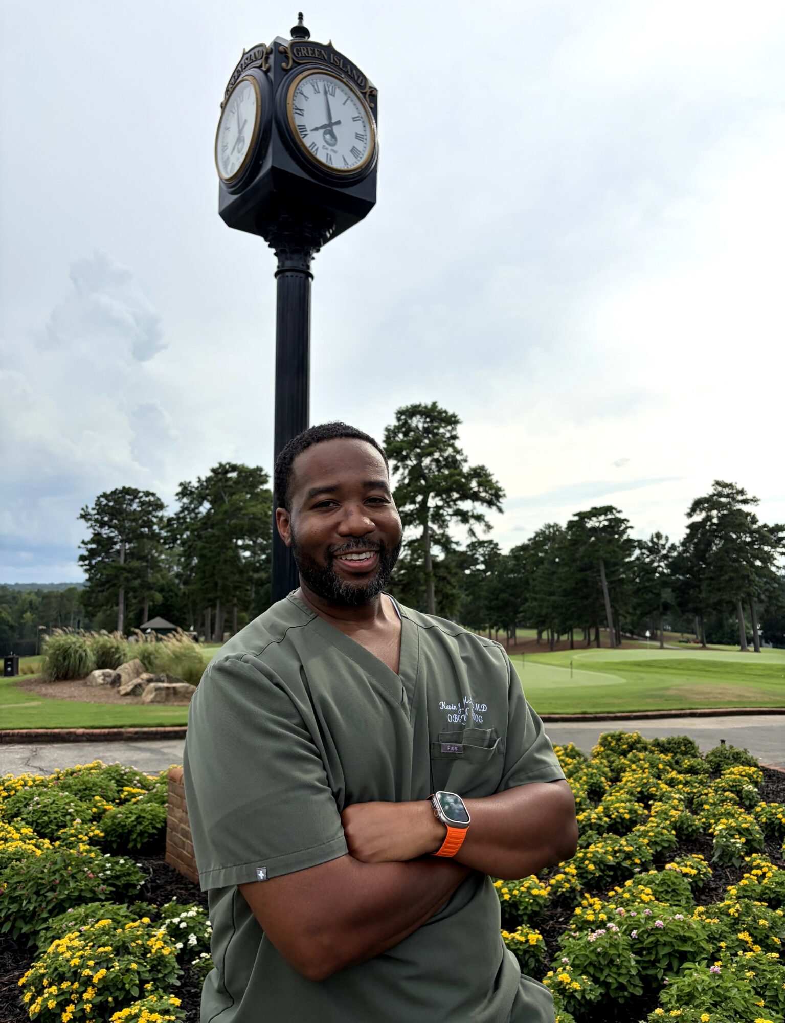 Man with arms crossed smiling outdoors near a clock tower and green landscape.