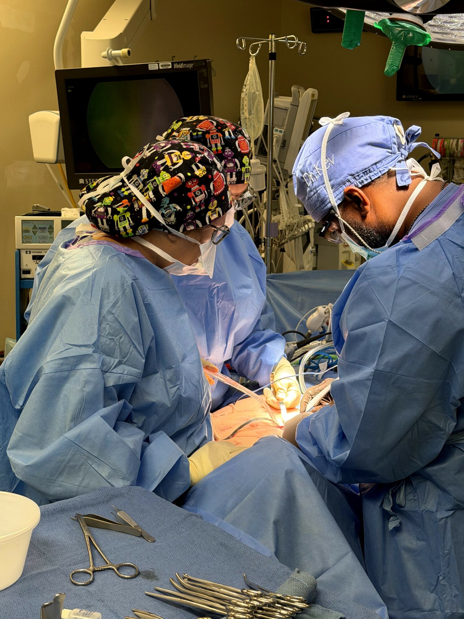 Two medical professionals in surgical attire perform a procedure in an operating room, with medical equipment and monitors nearby.