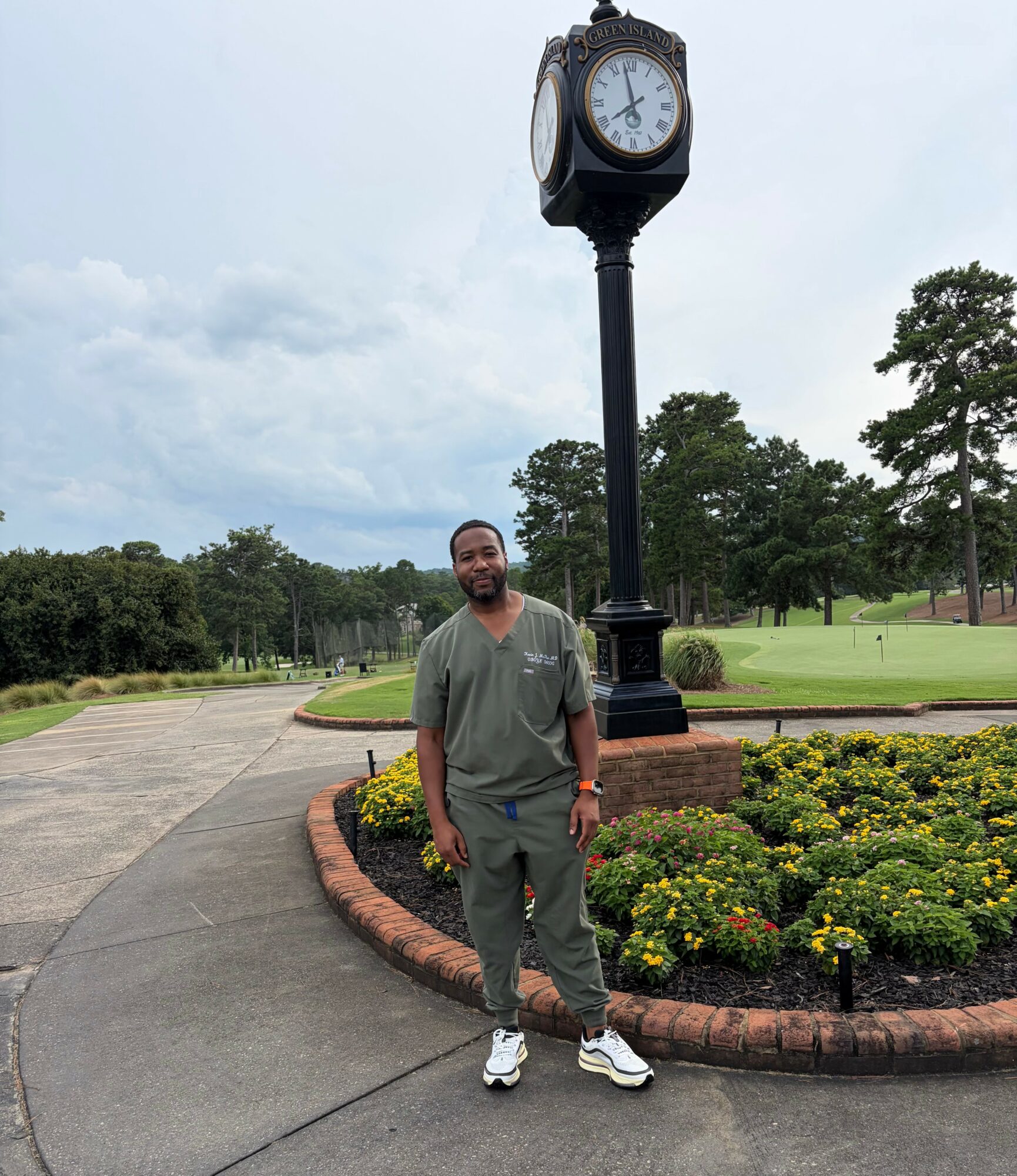 Man standing outdoors near a flower bed and a tall clock on a pole, with trees and cloudy sky in background.