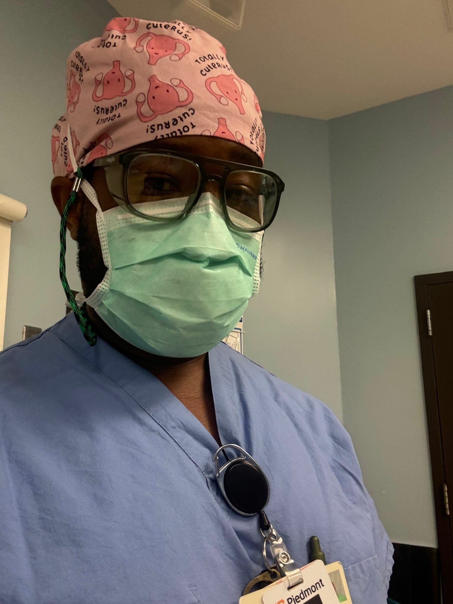 Person wearing glasses, a pink headscarf, a green face mask, and blue scrubs, in a medical setting.
