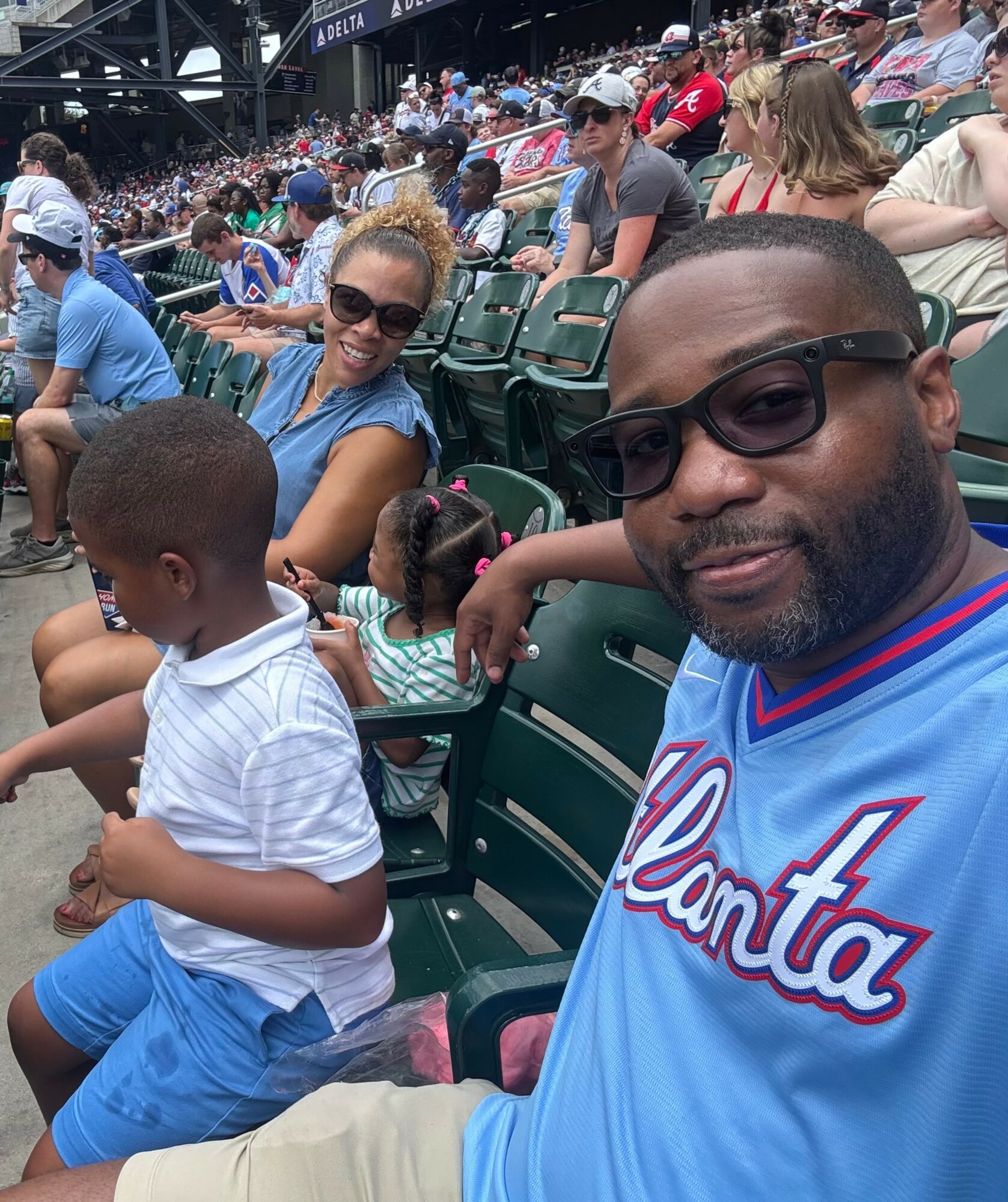 Man and woman with children sitting in stadium seats at a sports event, smiling, with many spectators in background.