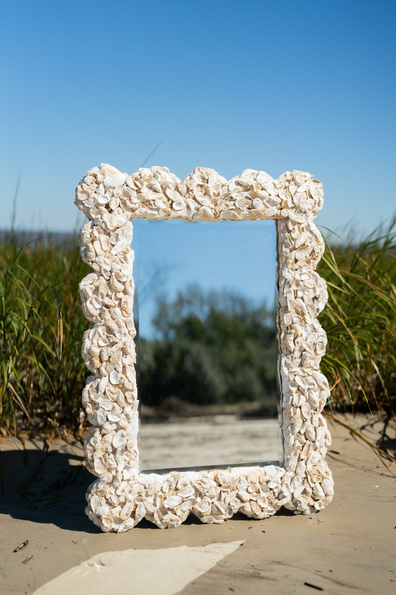 A rectangular mirror with a frame made of seashells, standing outdoors on sand with grass and blue sky in background.