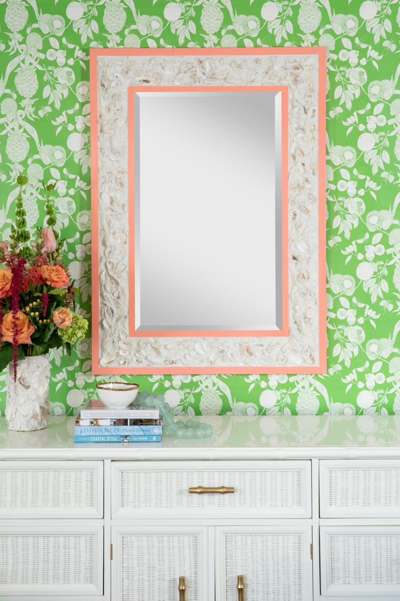 Decorative mirror with pink frame on green floral wallpaper, white dresser with books and flowers, and a bowl.