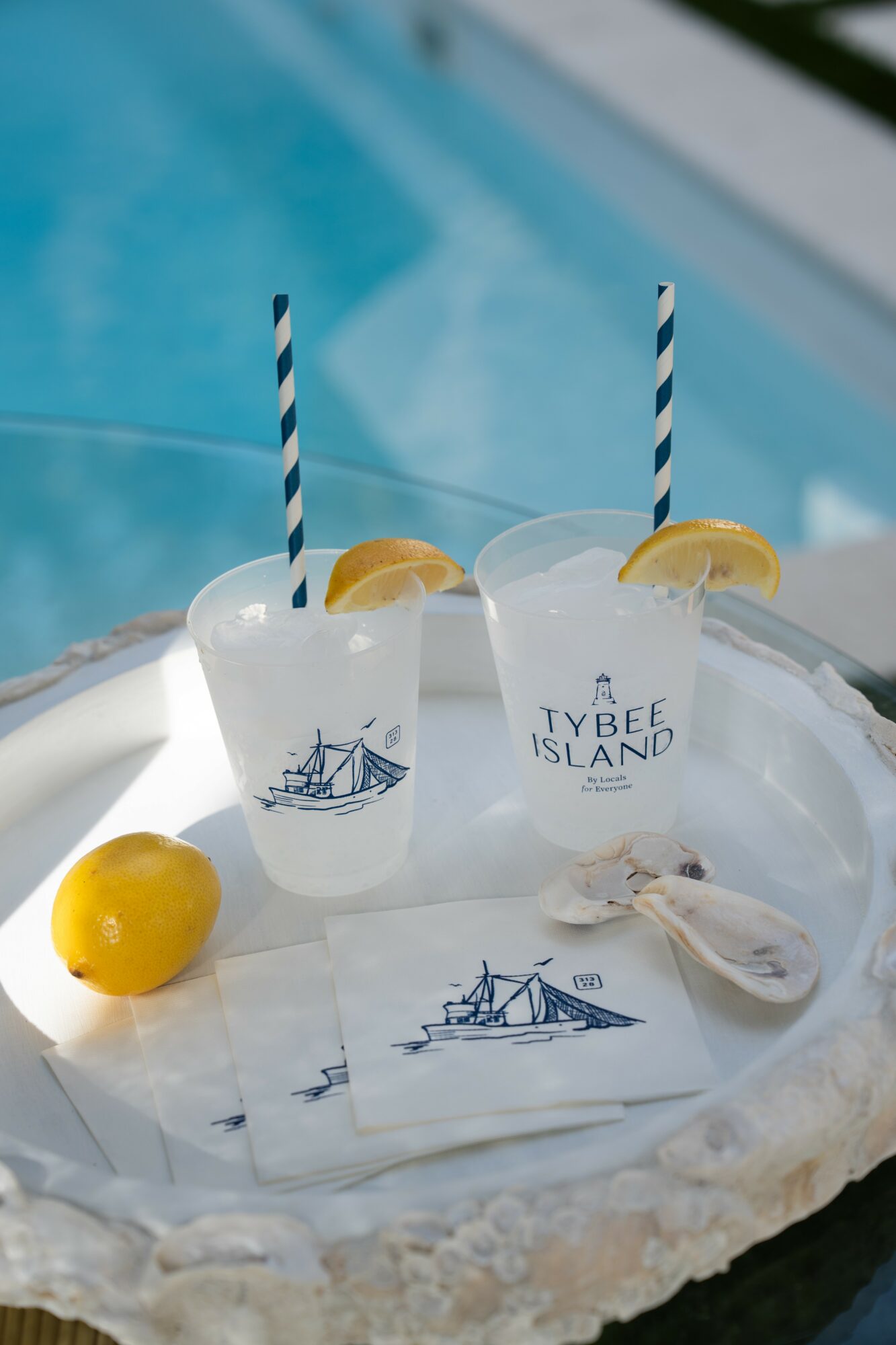 Two drinks with lemon slices and striped straws on a tray, a lemon, and a fish, with a pool in the background.
