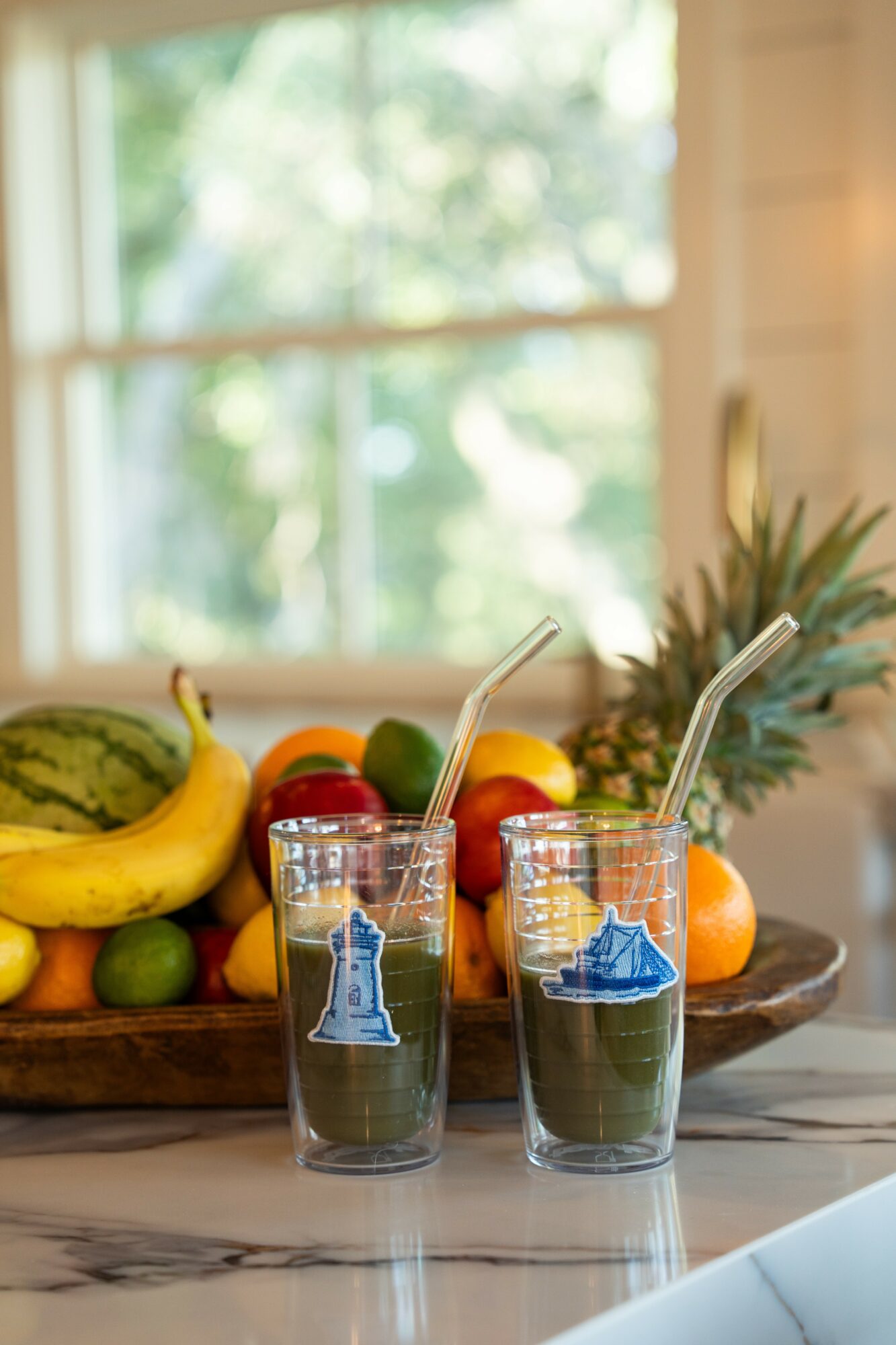 Two glasses with blue liquid and straws, a wooden tray of fruits including bananas, oranges, and limes, on a marble surface.