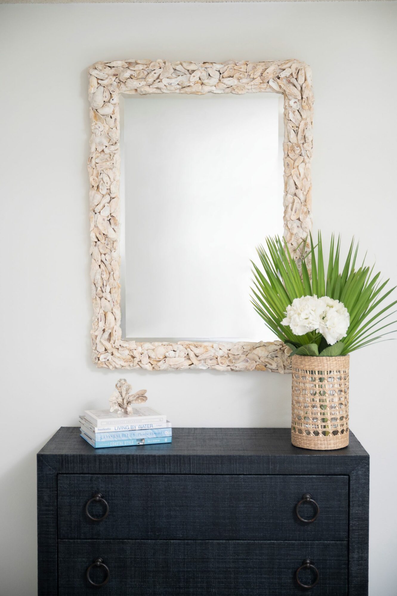 Decorative mirror with a textured frame hangs above a black dresser, with a vase of white flowers and green leaves on top.