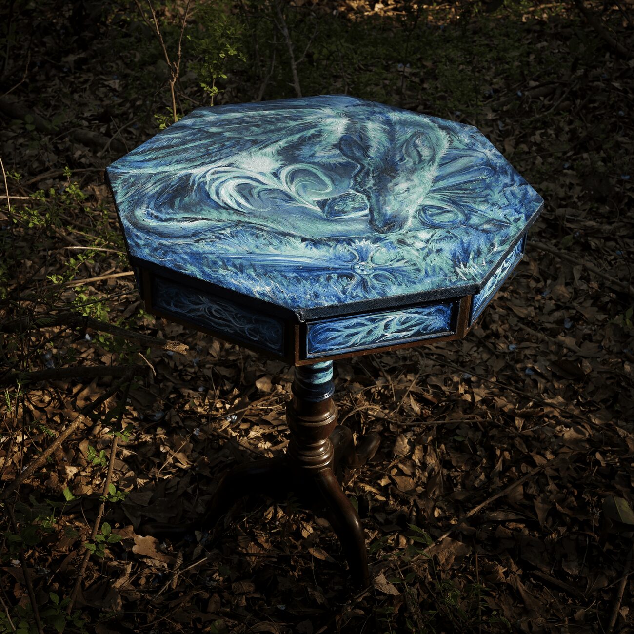 Octagonal table with a swirling blue and white marble pattern top, standing on a carved wooden pedestal base in a forest.