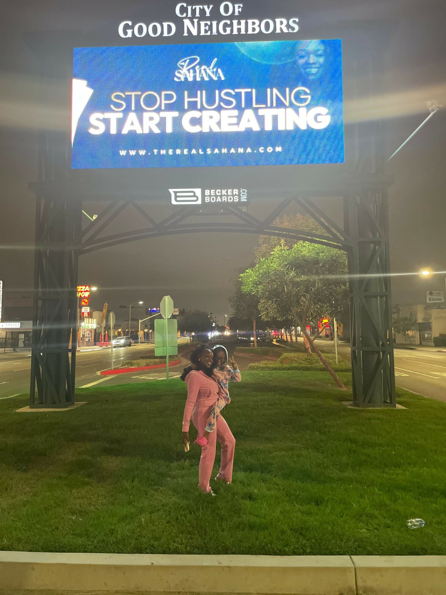 Nighttime city street with a digital sign, trees, and a person walking on grass, illuminated by streetlights.