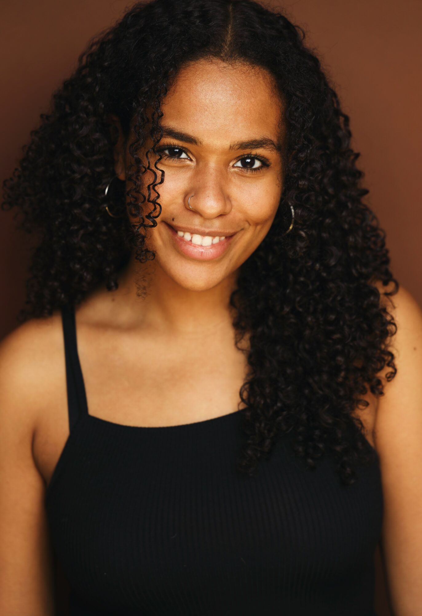 Young woman with curly hair smiling, wearing a black tank top, against a brown background.