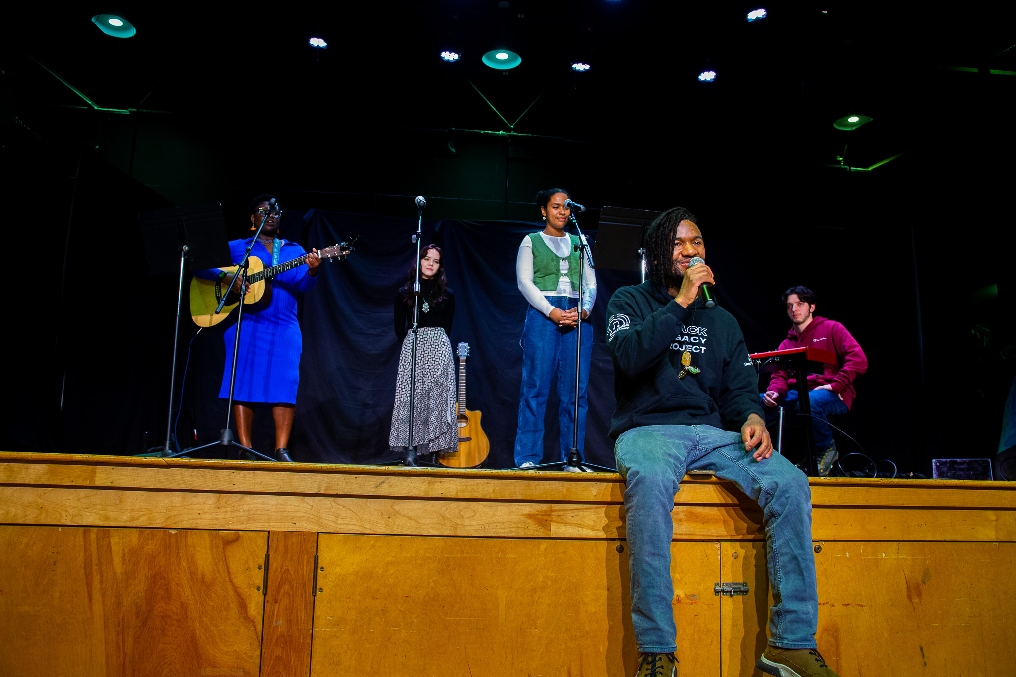 Five people on stage, one sitting and four standing, with musical instruments and microphones, in a performance setting.