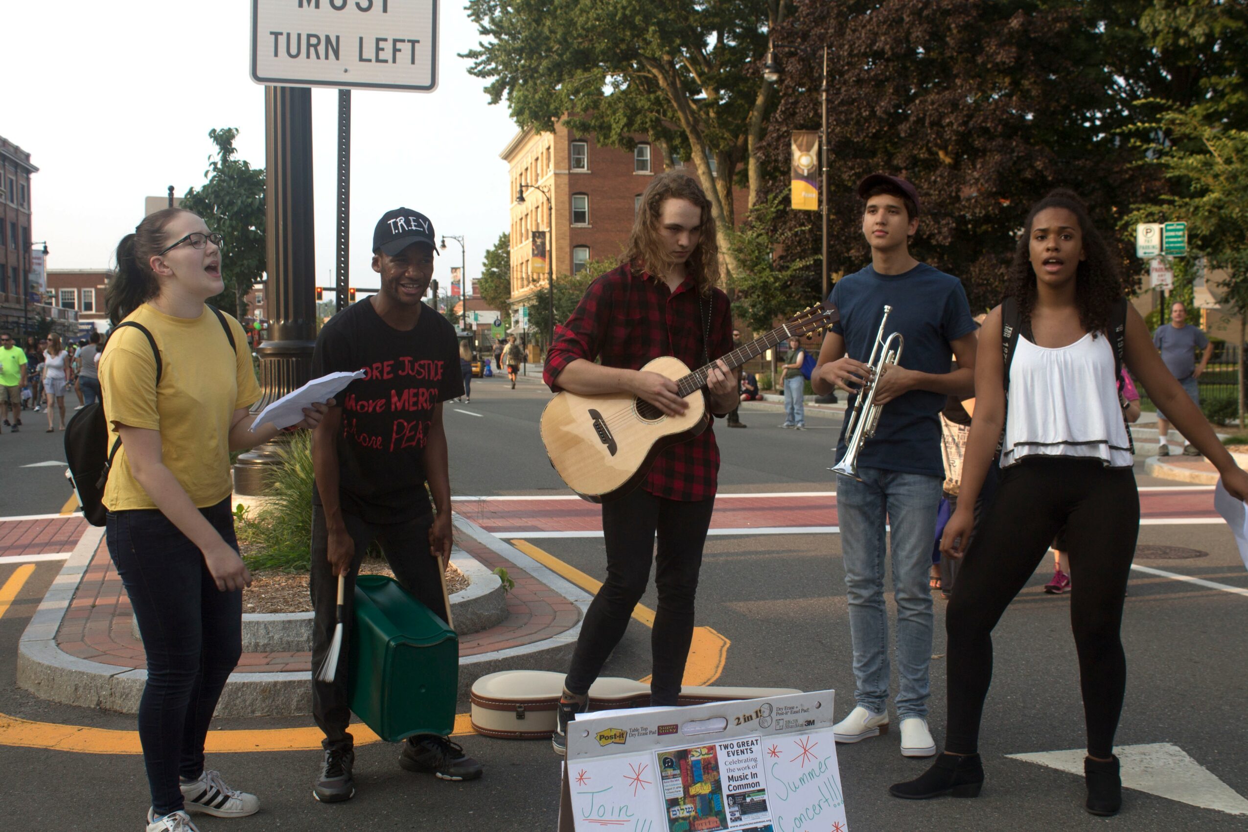 Group of five young people performing music on a city street corner, with trees and buildings in the background.