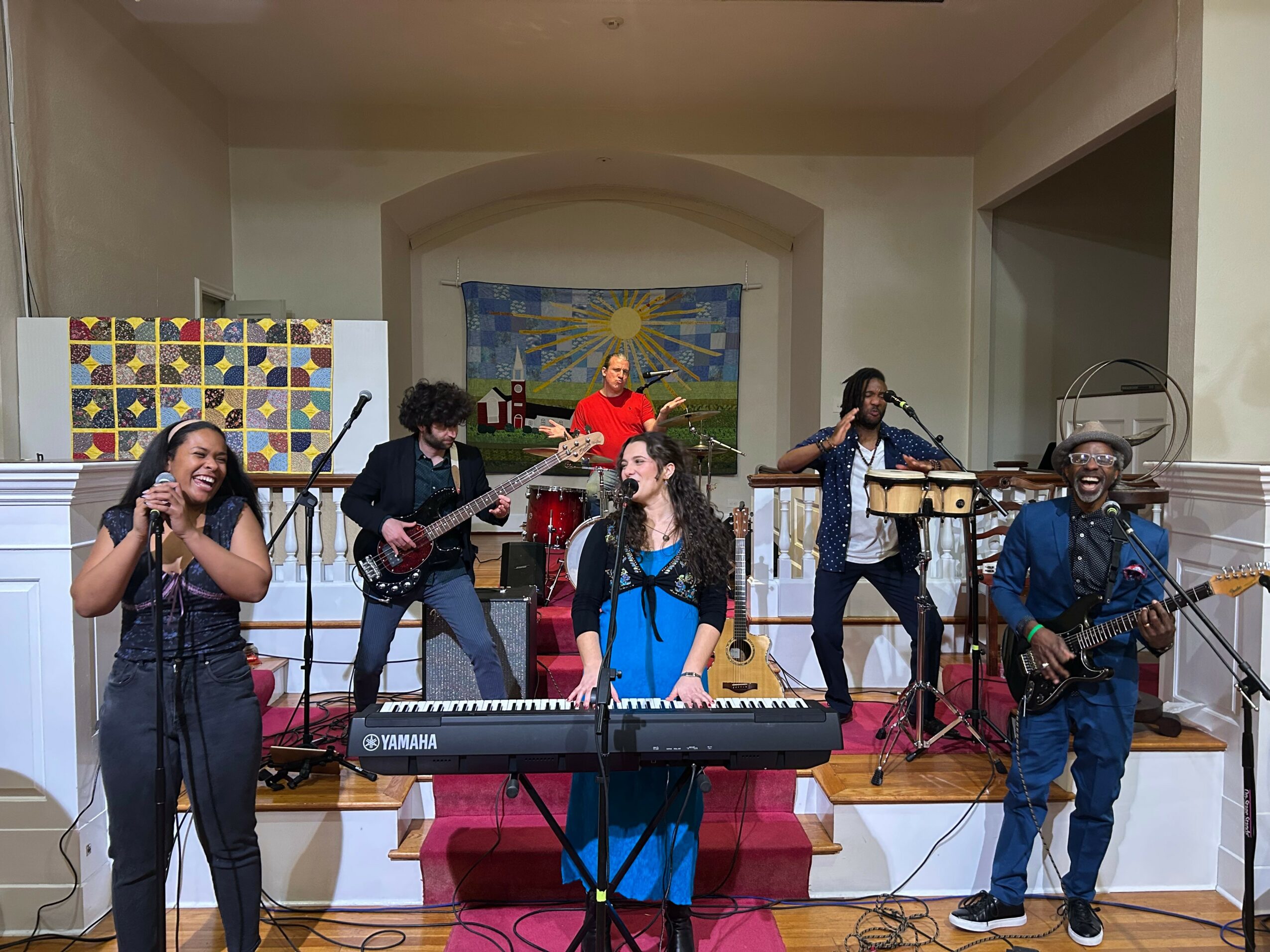 Group of six musicians performing on stage with instruments, including keyboard, guitars, and drums, in a room with colorful artwork.