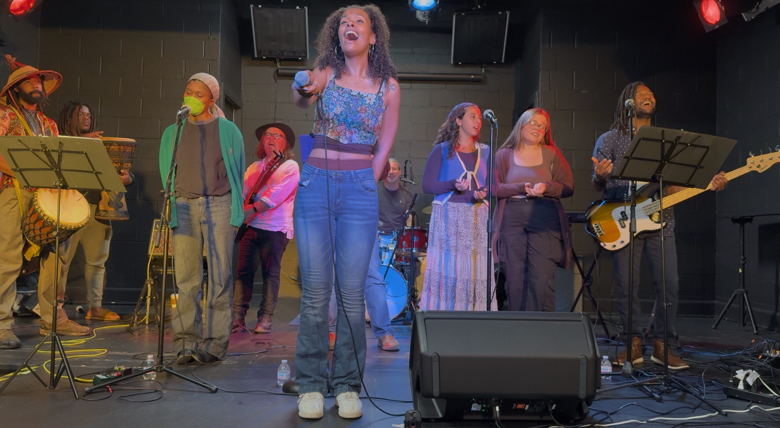 Group of musicians performing on stage, with a woman singing in the center, others playing instruments and singing, stage lights overhead.