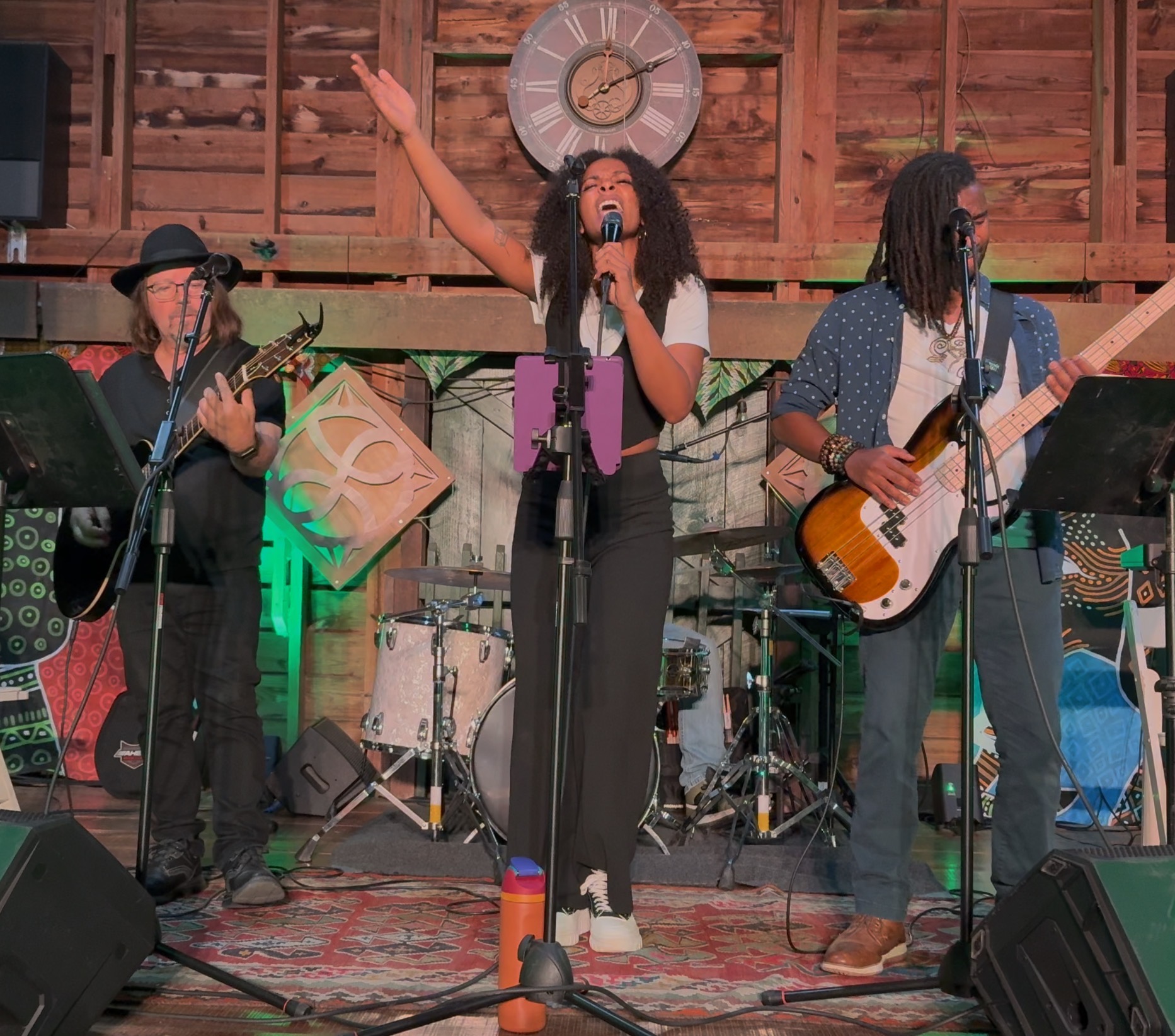 Three musicians perform on stage with instruments, one singing with hand raised, wooden backdrop, clock, and colorful decorations.