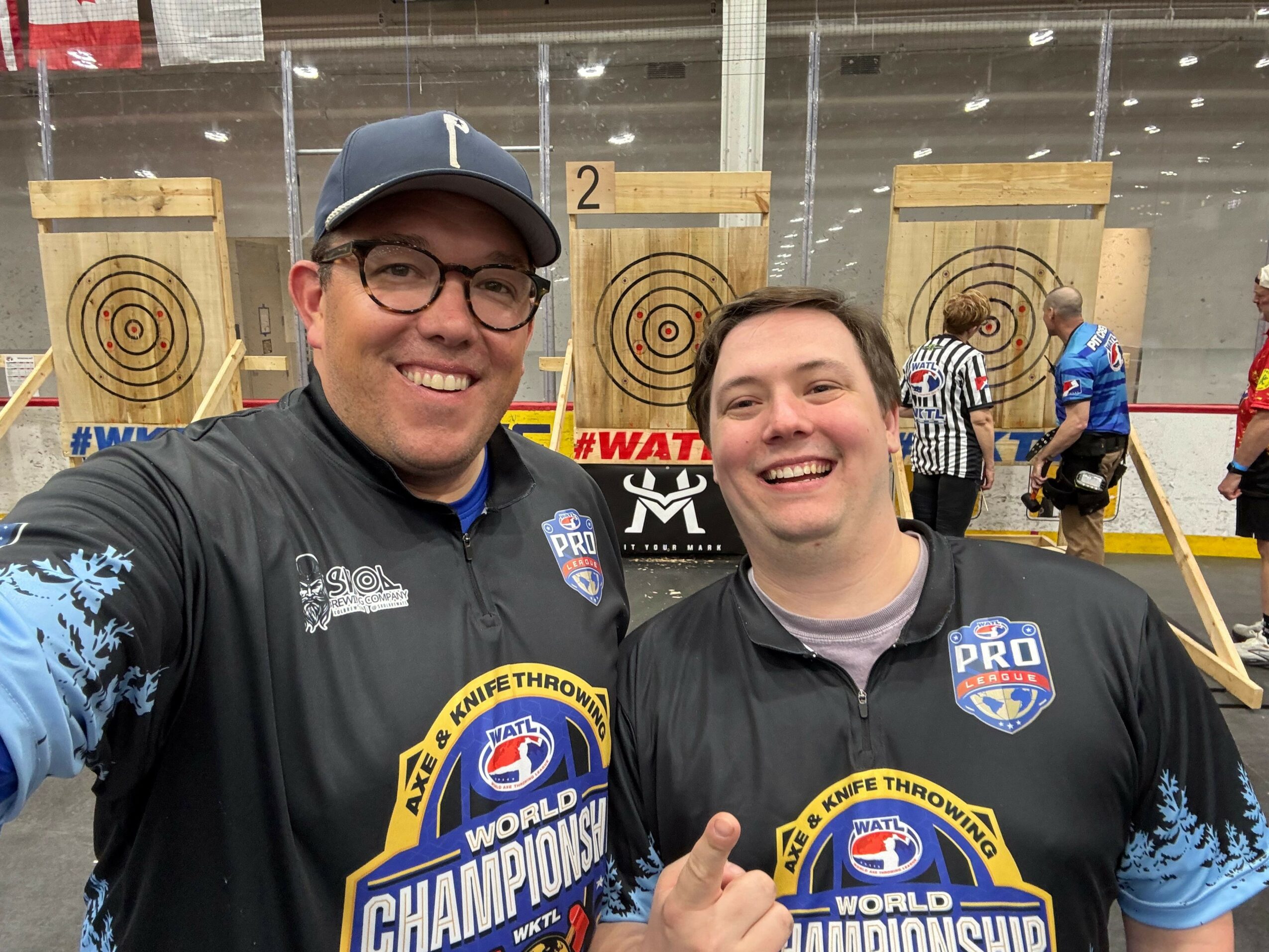 Two people smiling at an indoor archery range with targets behind them, one wearing glasses and a cap, both in black shirts.