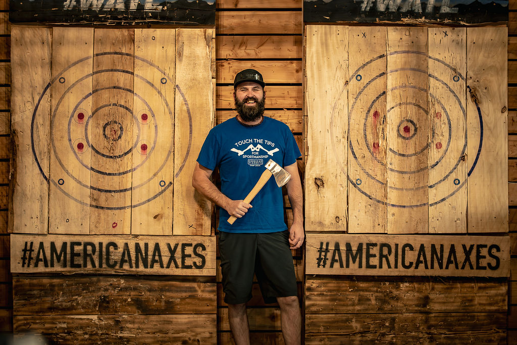 Man with a beard holding a wooden axe, standing in front of two wooden axe targets with hashtag #AMERICANAxes below, outdoors.