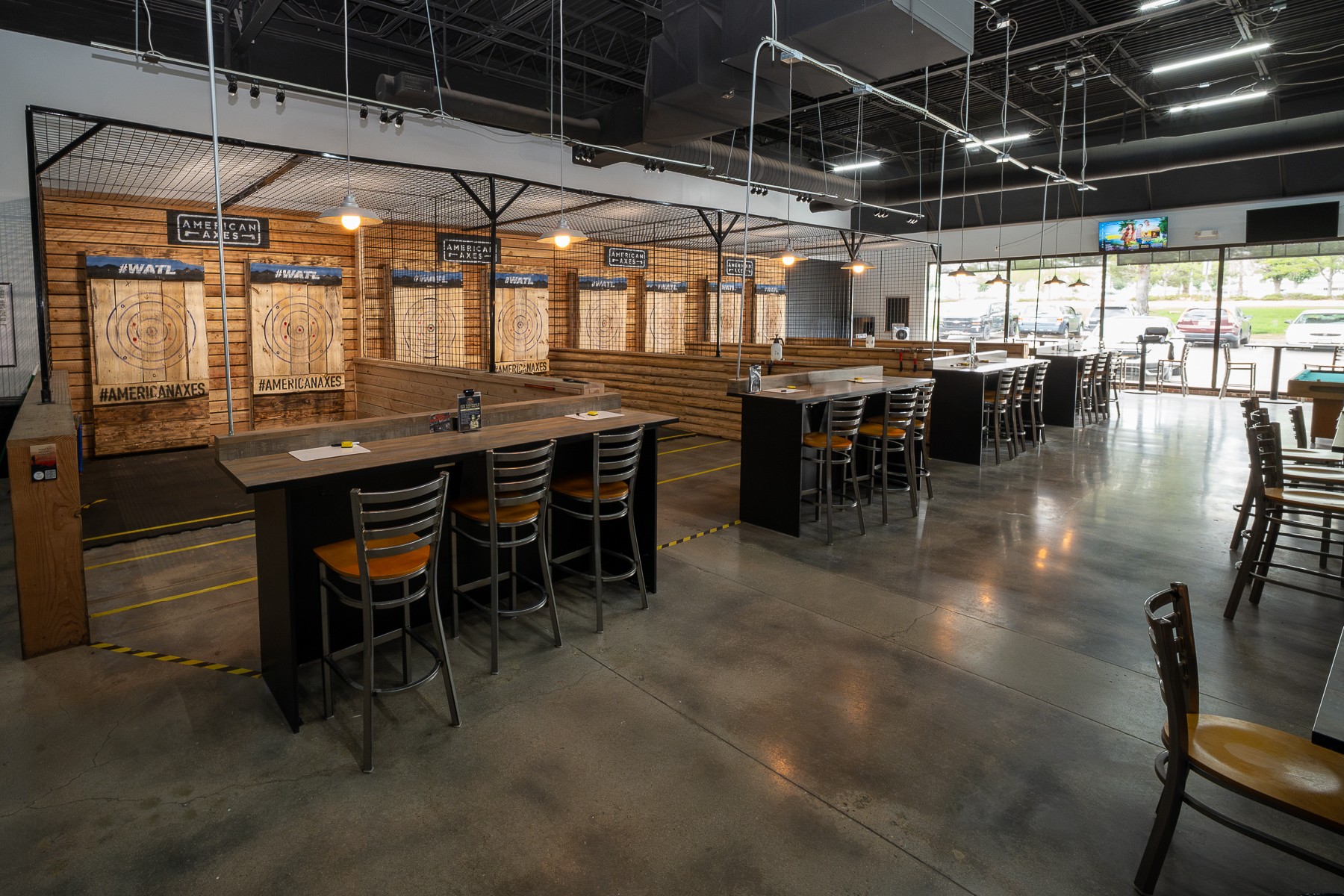 Interior of a modern restaurant or bar with high tables, chairs, and a brick wall with wooden panels, large windows, and polished concrete floor.