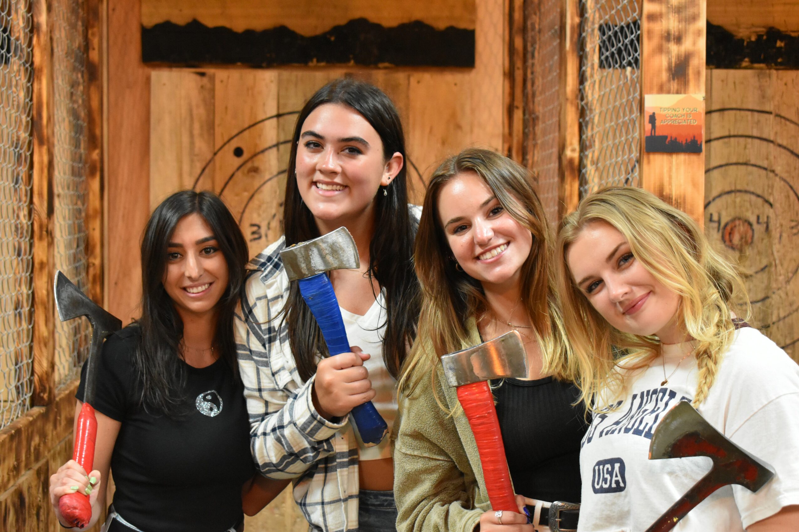 Four women smiling, holding axes, standing indoors with wooden walls and targets in the background.