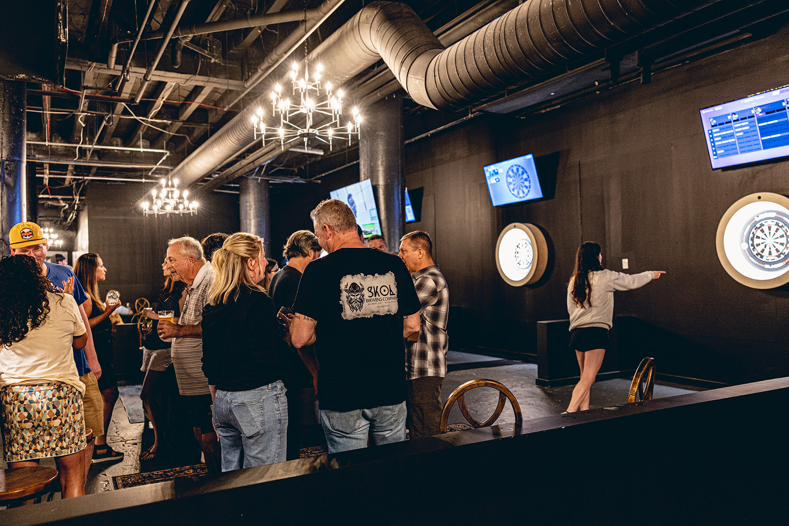 People gather in an indoor space with dartboards, screens, and industrial ceiling pipes, some playing darts and others watching.