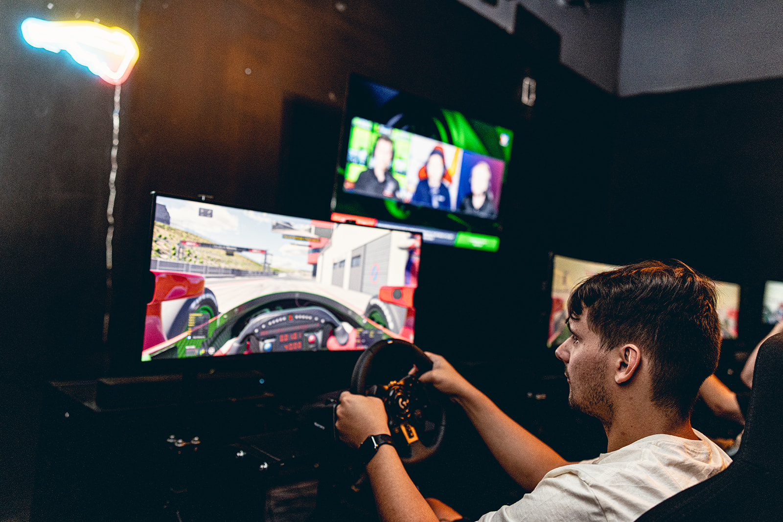 Person with short dark hair driving a racing simulator, multiple screens displaying racing scenes in the background.