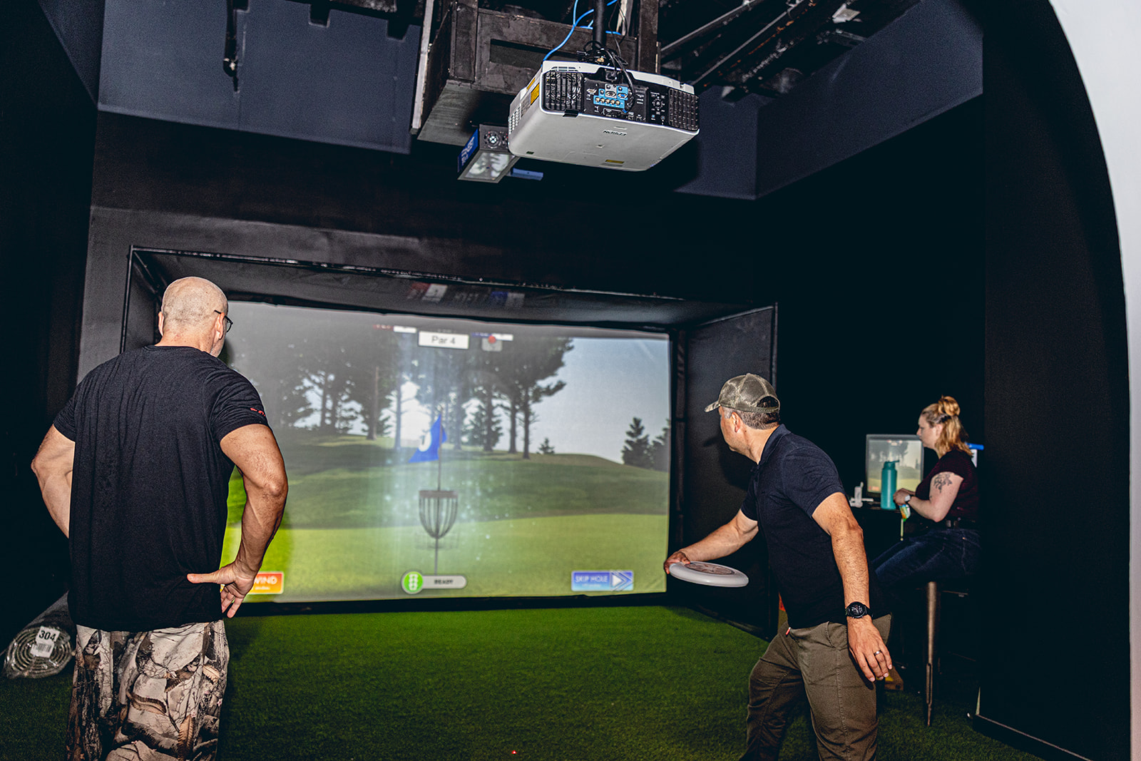 Three people playing indoor golf on a large screen, with one person preparing to take a shot, in a dark room.