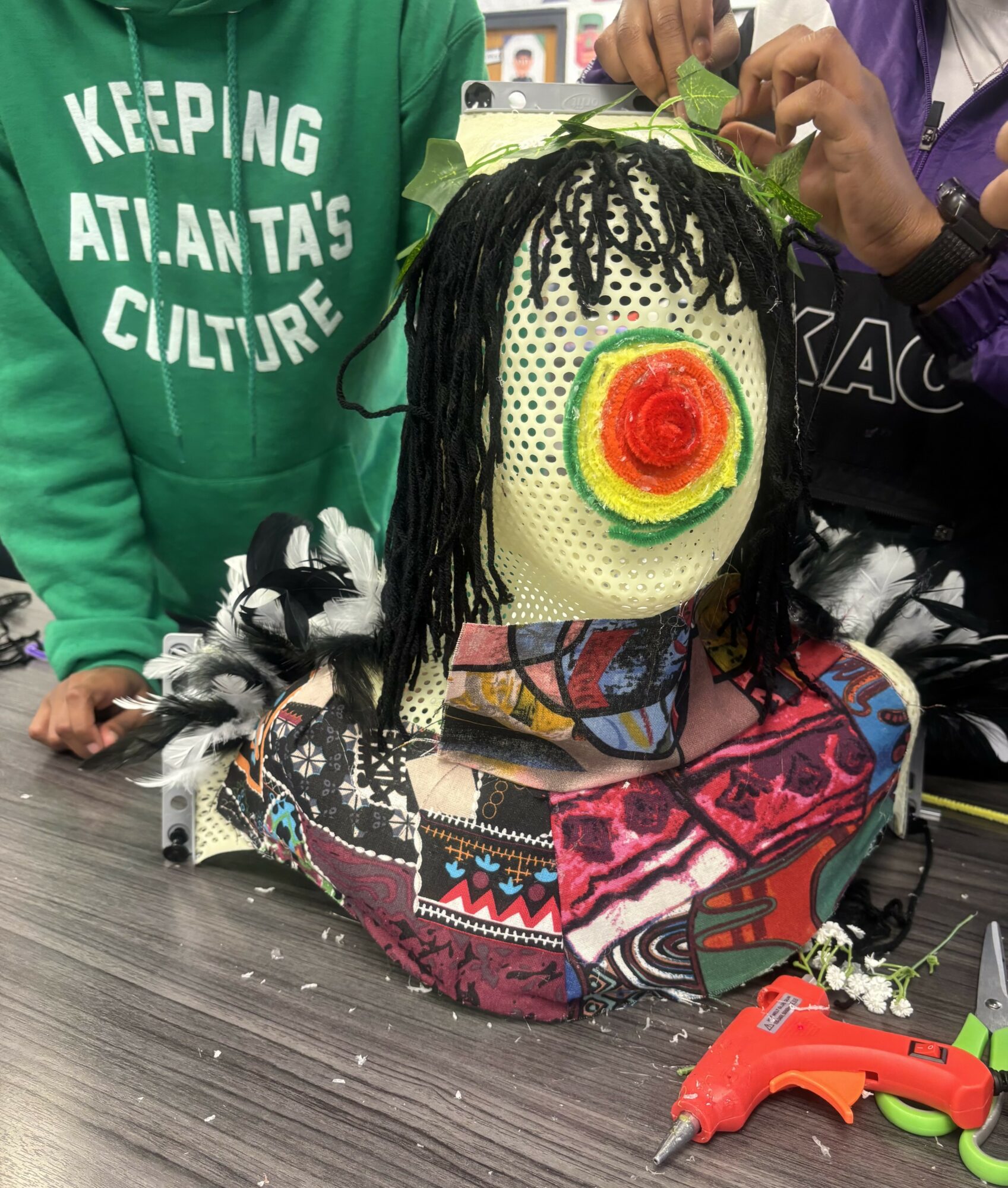 Children's hands adjusting a clown mask with a red, yellow, and green spiral face, on a colorful bag, on a table.