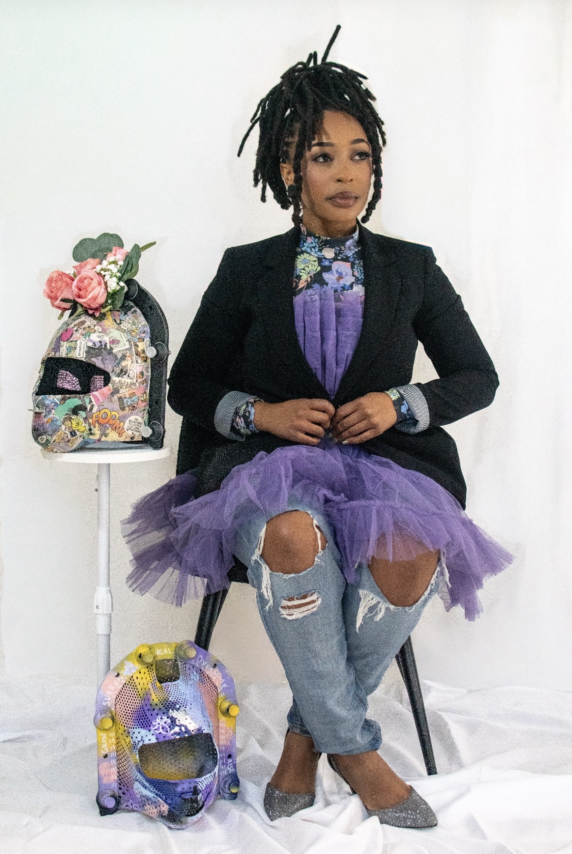 Woman with dreadlocks wearing a black blazer, purple tulle skirt, and ripped jeans, sitting with a backpack and flowers.