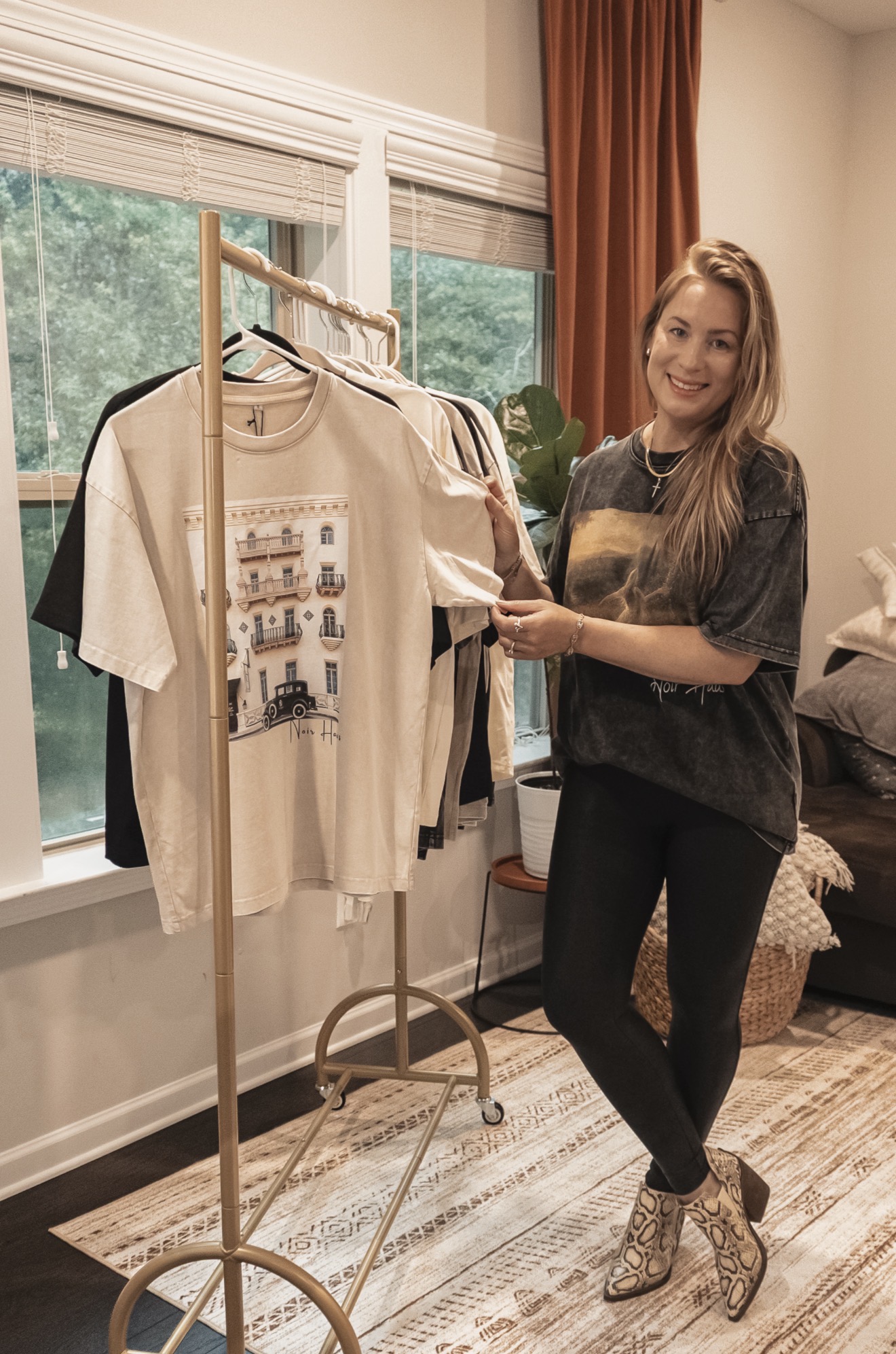 Woman with long hair smiling, holding a beige T-shirt with a graphic, standing next to a clothing rack inside a room.