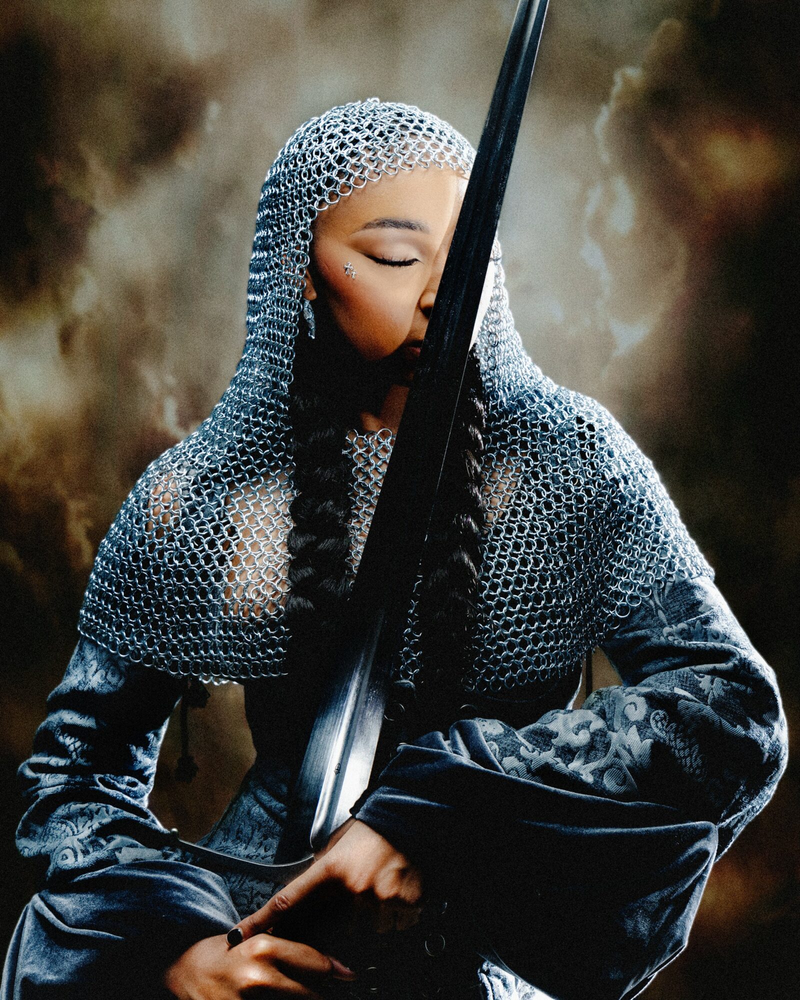 A woman in chainmail armor holds a sword vertically, with her eyes closed, in a dark, smoky background.