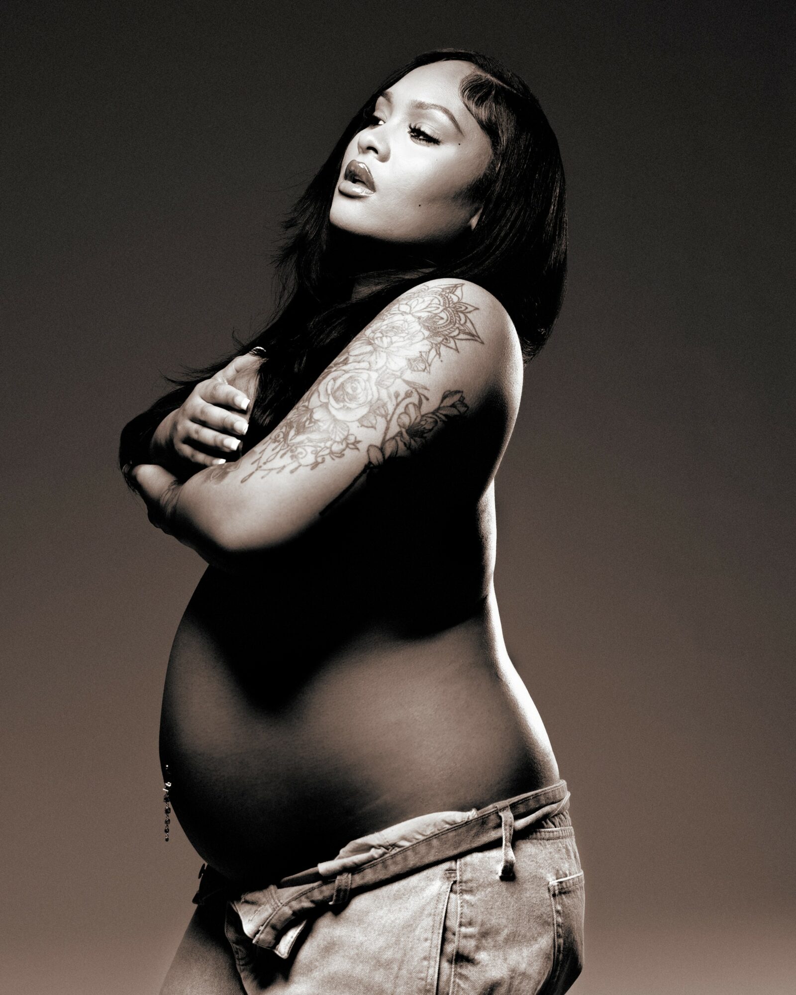 Pregnant woman with long dark hair and tattoos on her arm, posing with arms crossed, in black and white.