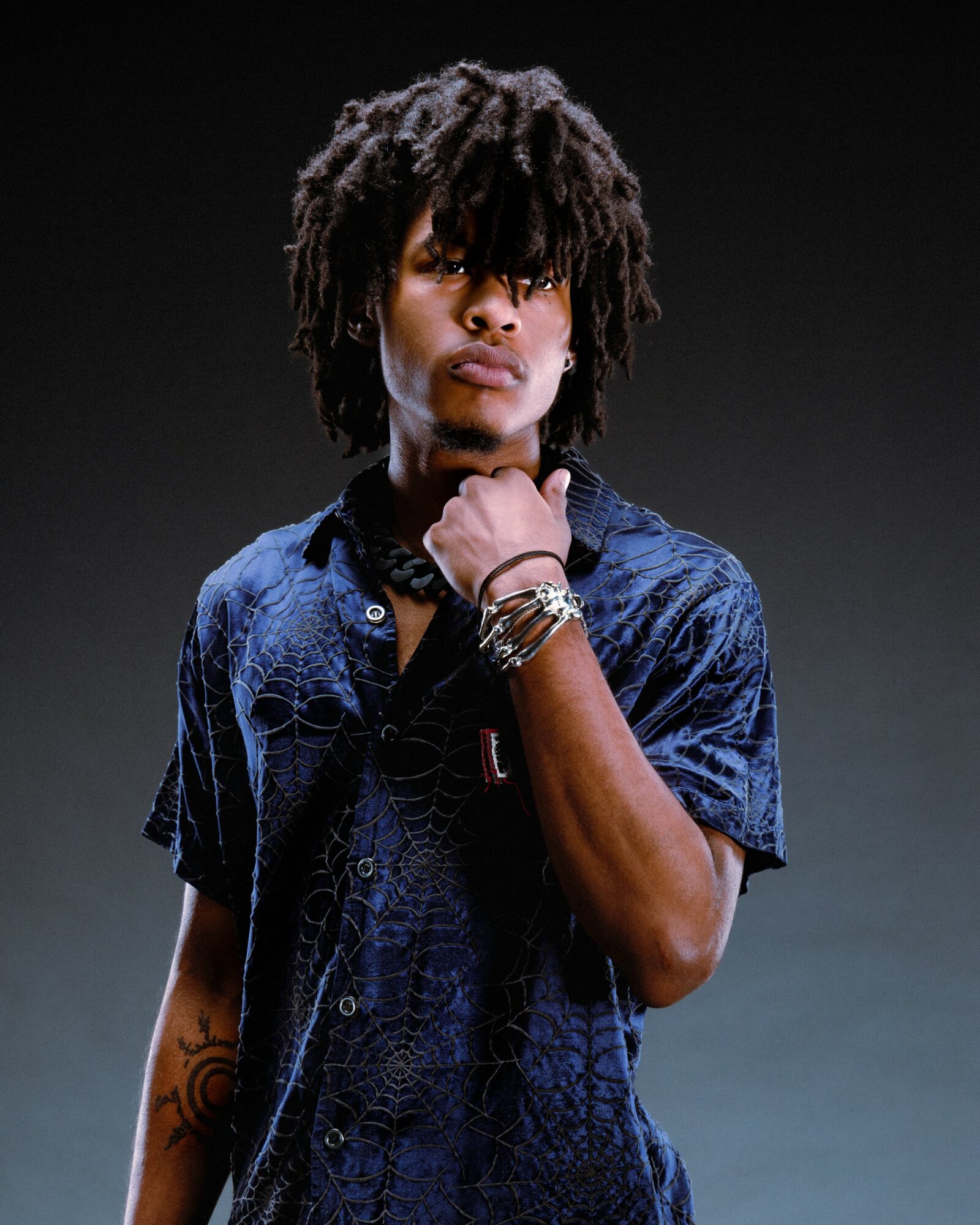 Young person with dreadlocks wearing a blue shirt, standing against a dark background, looking at the camera with a serious expression.