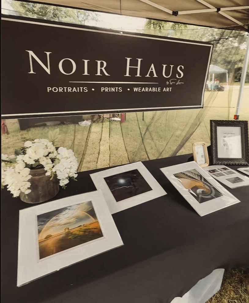 Display table with framed photographs, a flower arrangement, and a sign reading 'Noir Haus' at an outdoor event.