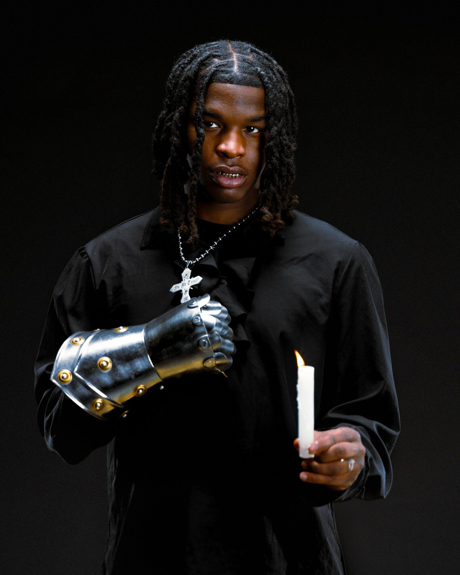 Young man with dreadlocks wearing a black shirt, a silver chain, and a metallic glove, holding a white object, against a black background.
