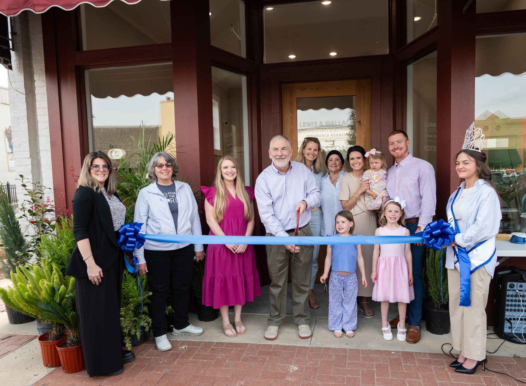 Group of people standing outside a building holding a ribbon for a ribbon-cutting event, with children and adults smiling.