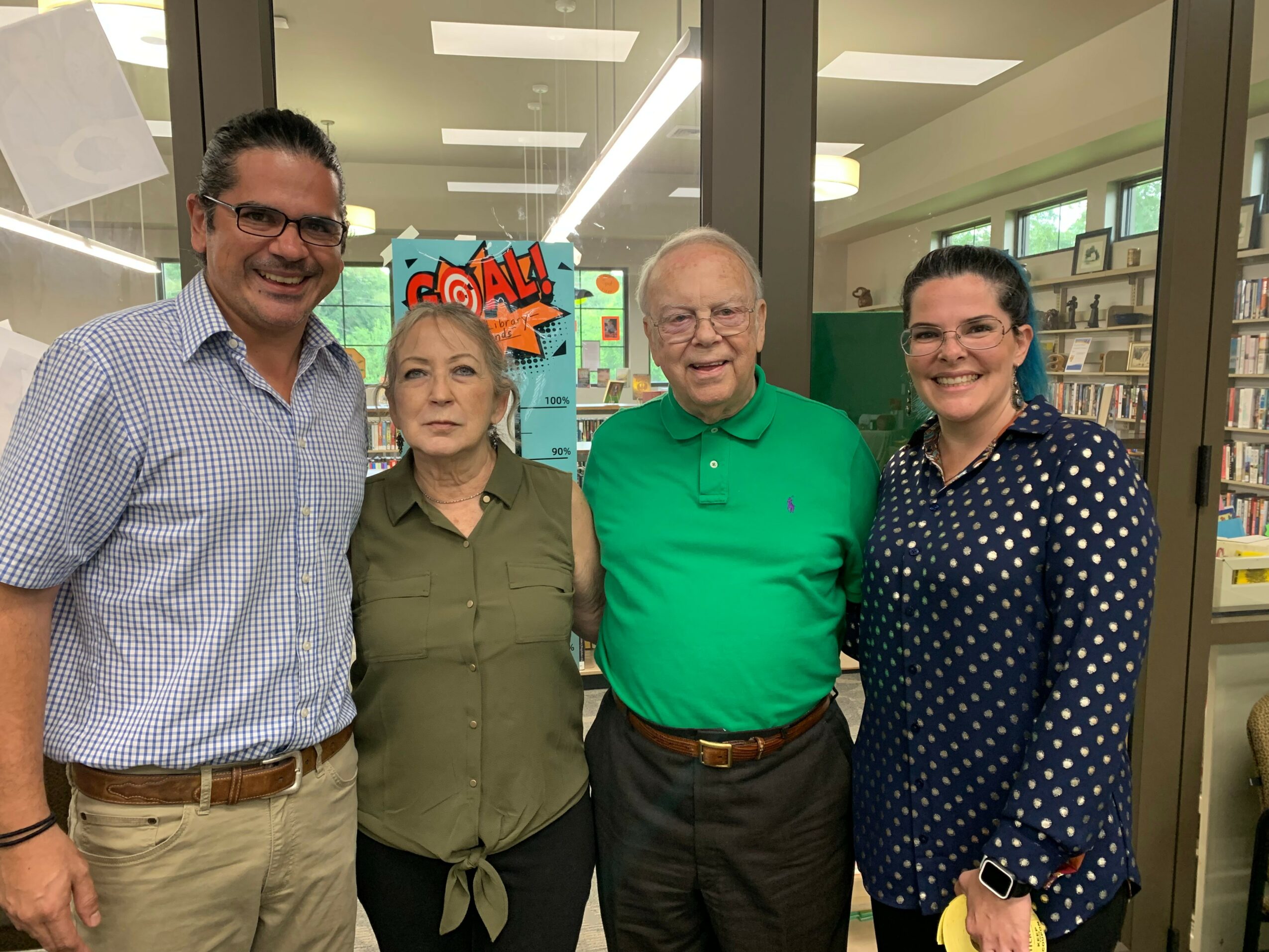 Four people standing together indoors, smiling, with bookshelves and a colorful sign in the background.