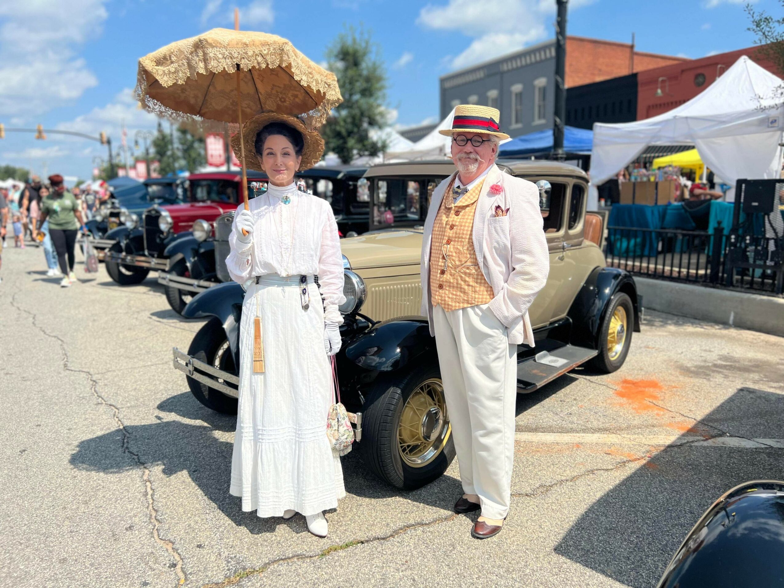 Two people dressed in vintage clothing stand beside a classic car at an outdoor event, with tents and people in the background.
