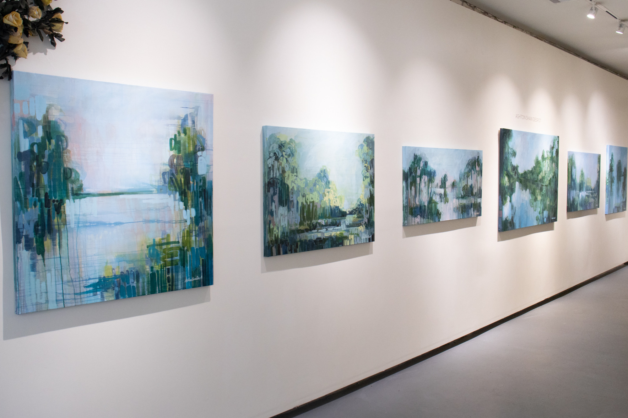 Gallery wall with multiple abstract landscape paintings displayed in a row on a white wall.