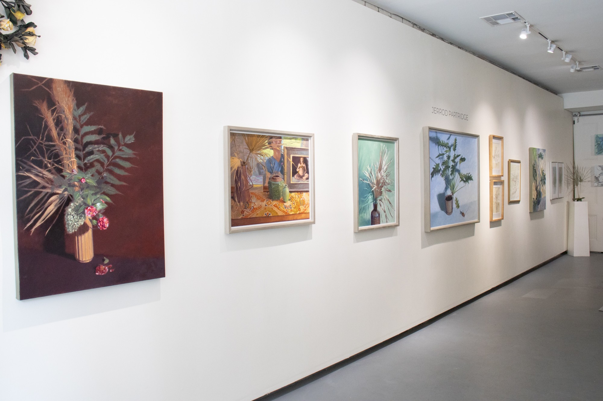 Gallery wall with various framed paintings and artworks displayed in a row, white wall background, ceiling lights, and a gray floor.